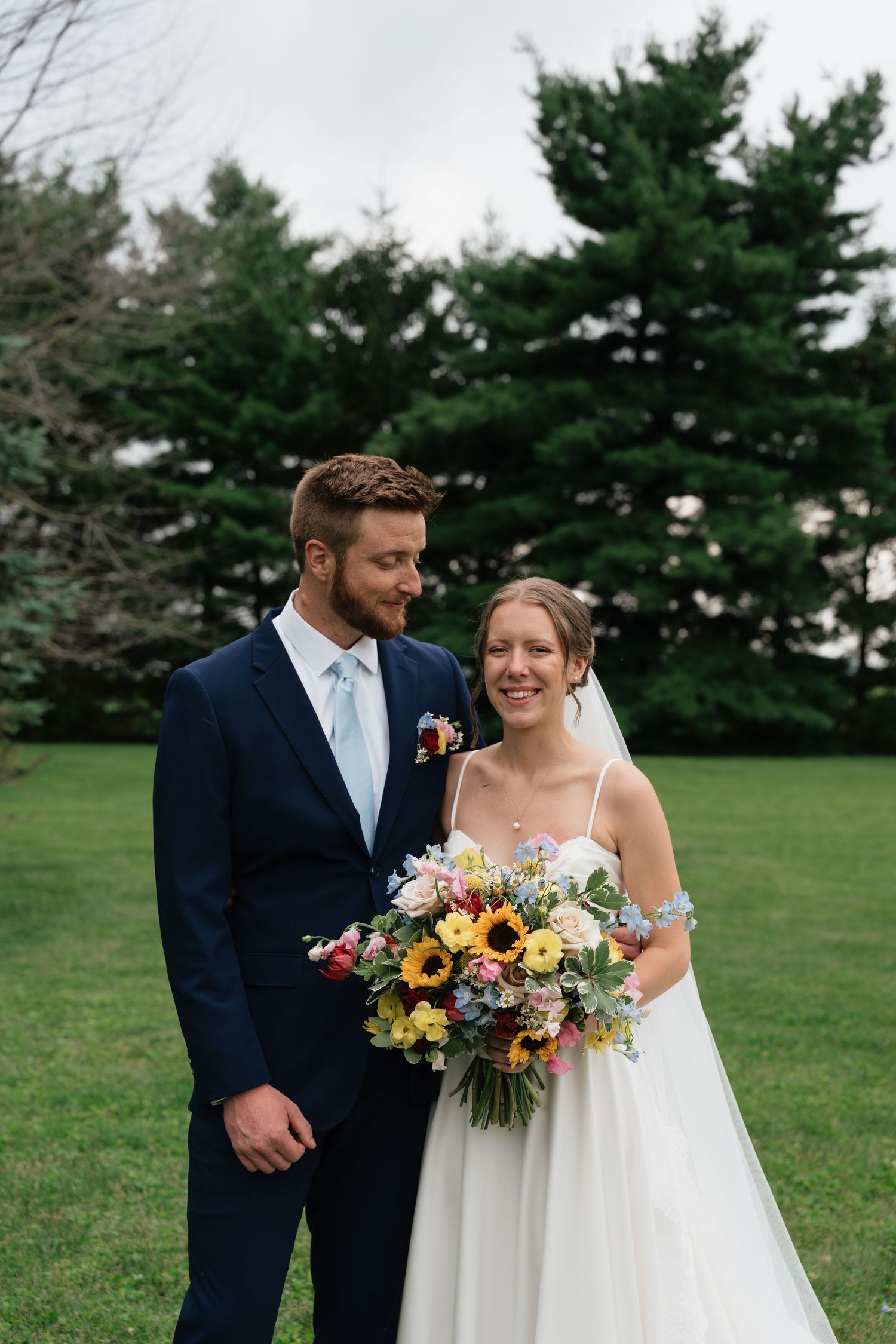 Bride holding colorful sunflower bouquet stands with groom in navy suit outdoors on green lawn with evergreen trees.