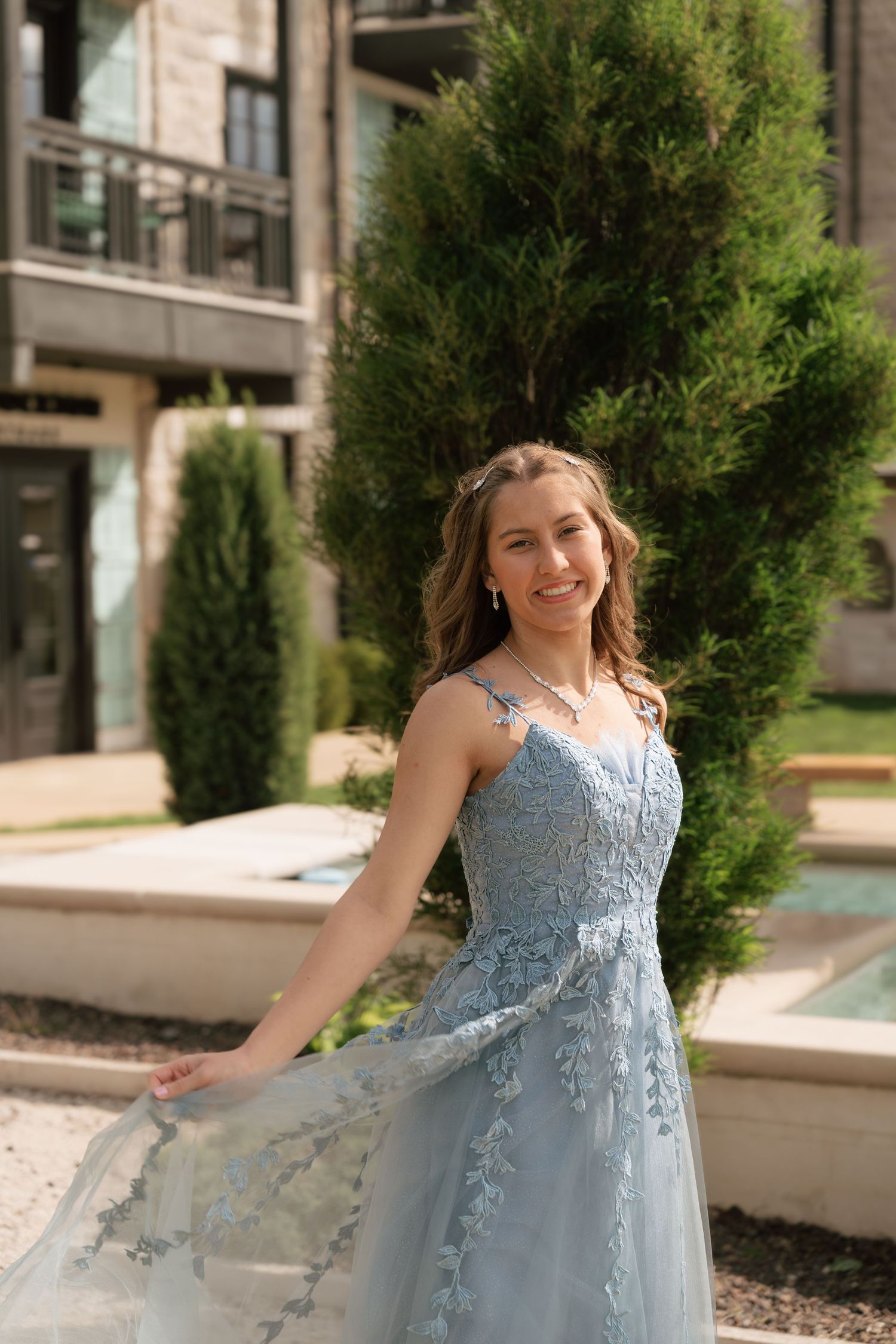 Young woman in a dusty blue floral lace A-line prom gown poses outdoors near evergreen trees and a fountain.