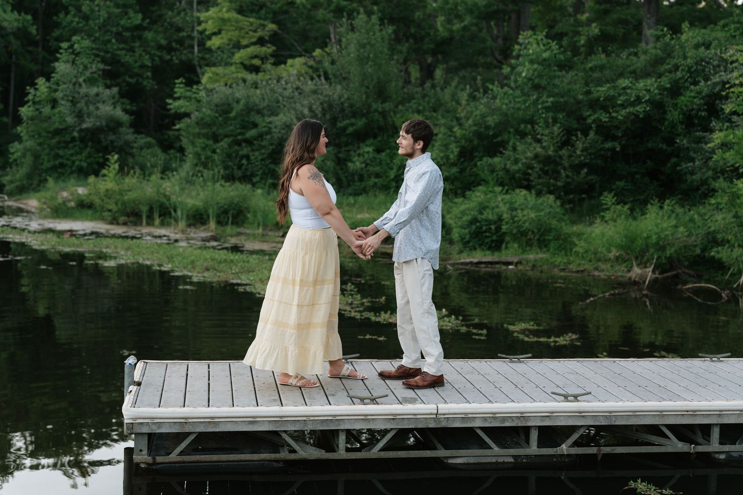 Couple holding hands on a wooden dock by a serene lake surrounded by lush green trees.