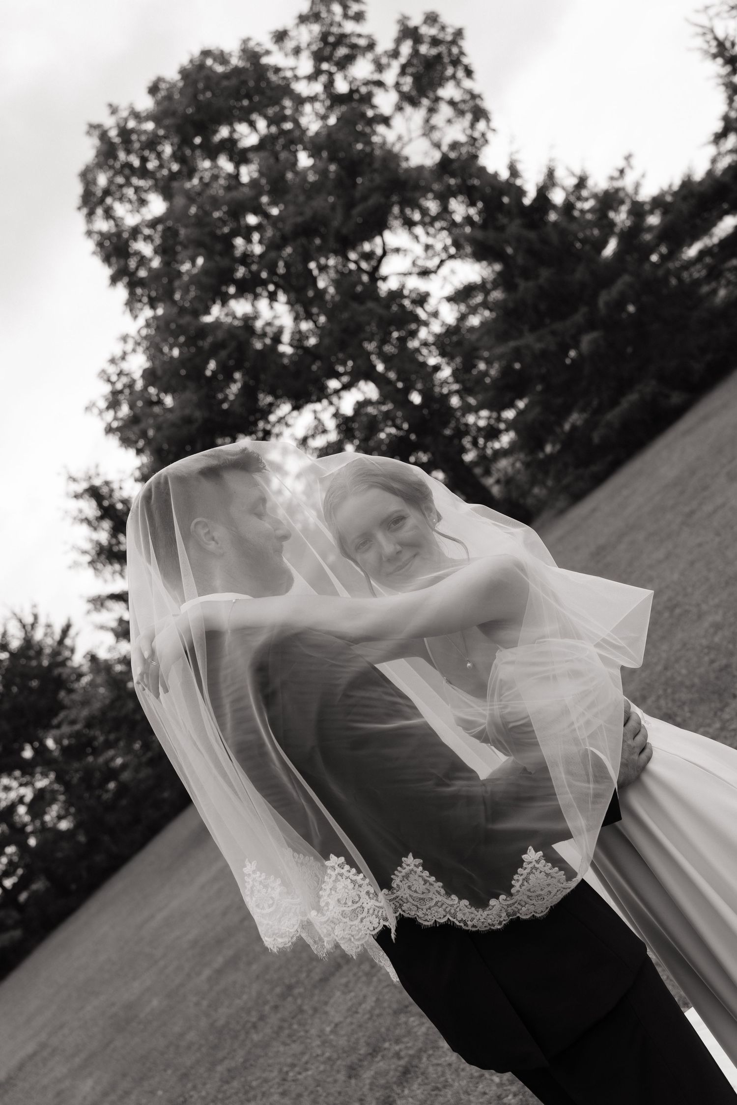 Black and white photo of bride and groom kissing under flowing veil outdoors on their wedding day.