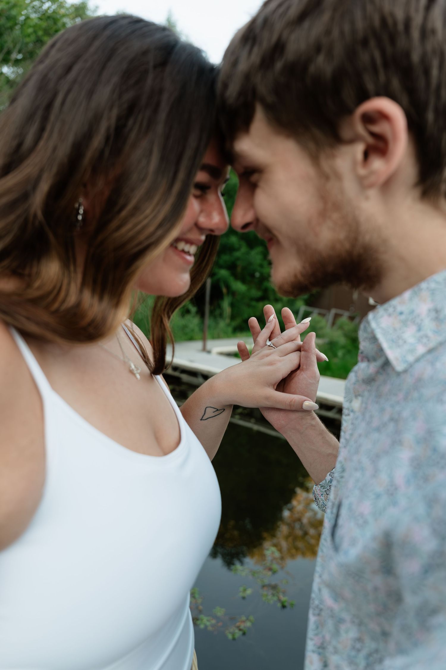 Happy couple touching foreheads and holding hands, showcasing an engagement ring near a peaceful pond.