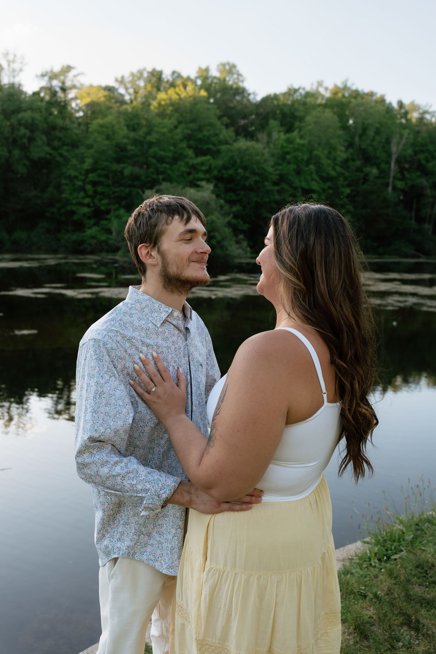 Couple embracing and smiling at each other by a serene lake surrounded by lush green trees at dusk.