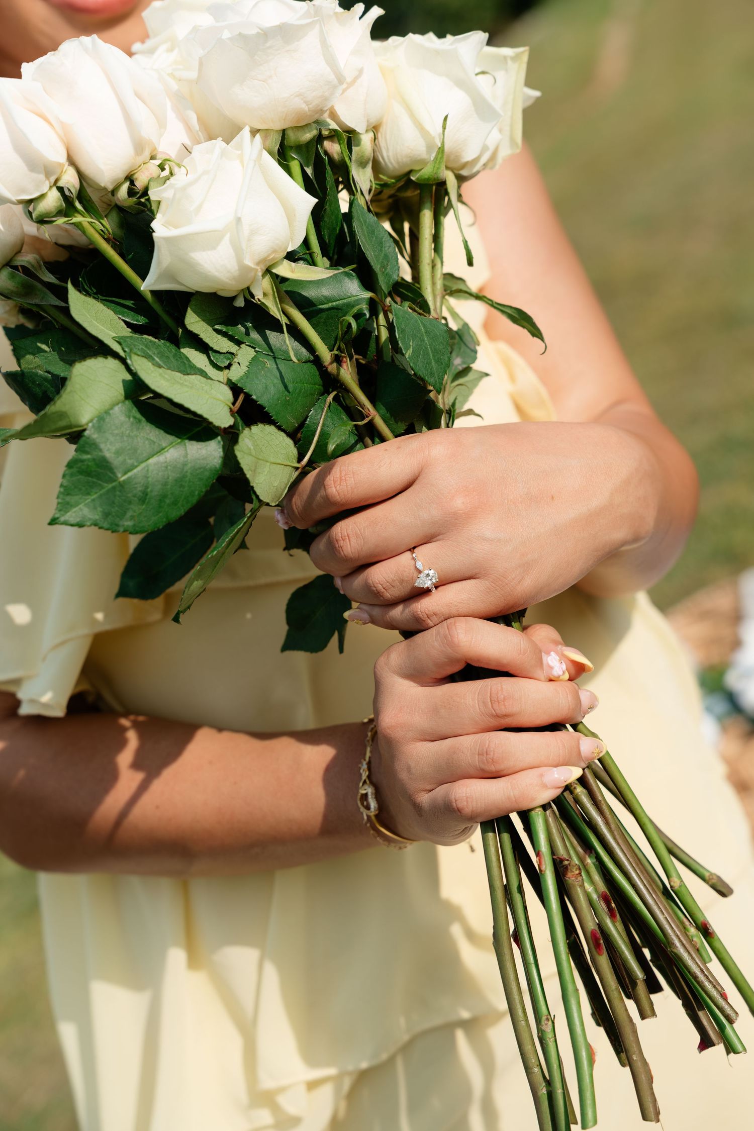 Bride holding white rose bouquet with diamond engagement ring and bracelet, wearing yellow dress outdoors.