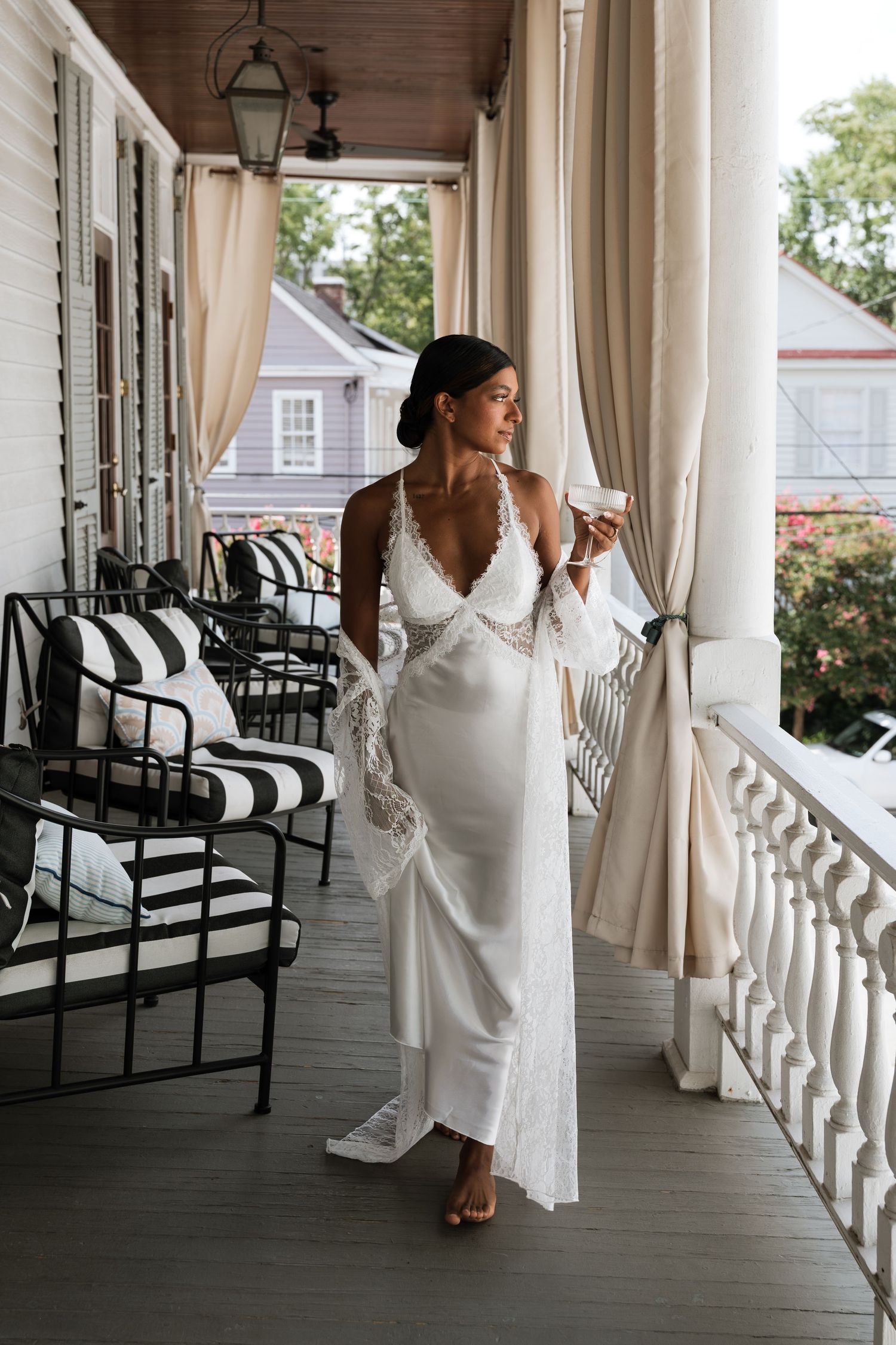 Woman in white satin slip dress holding coffee cup on elegant porch with striped chairs and cream curtains.