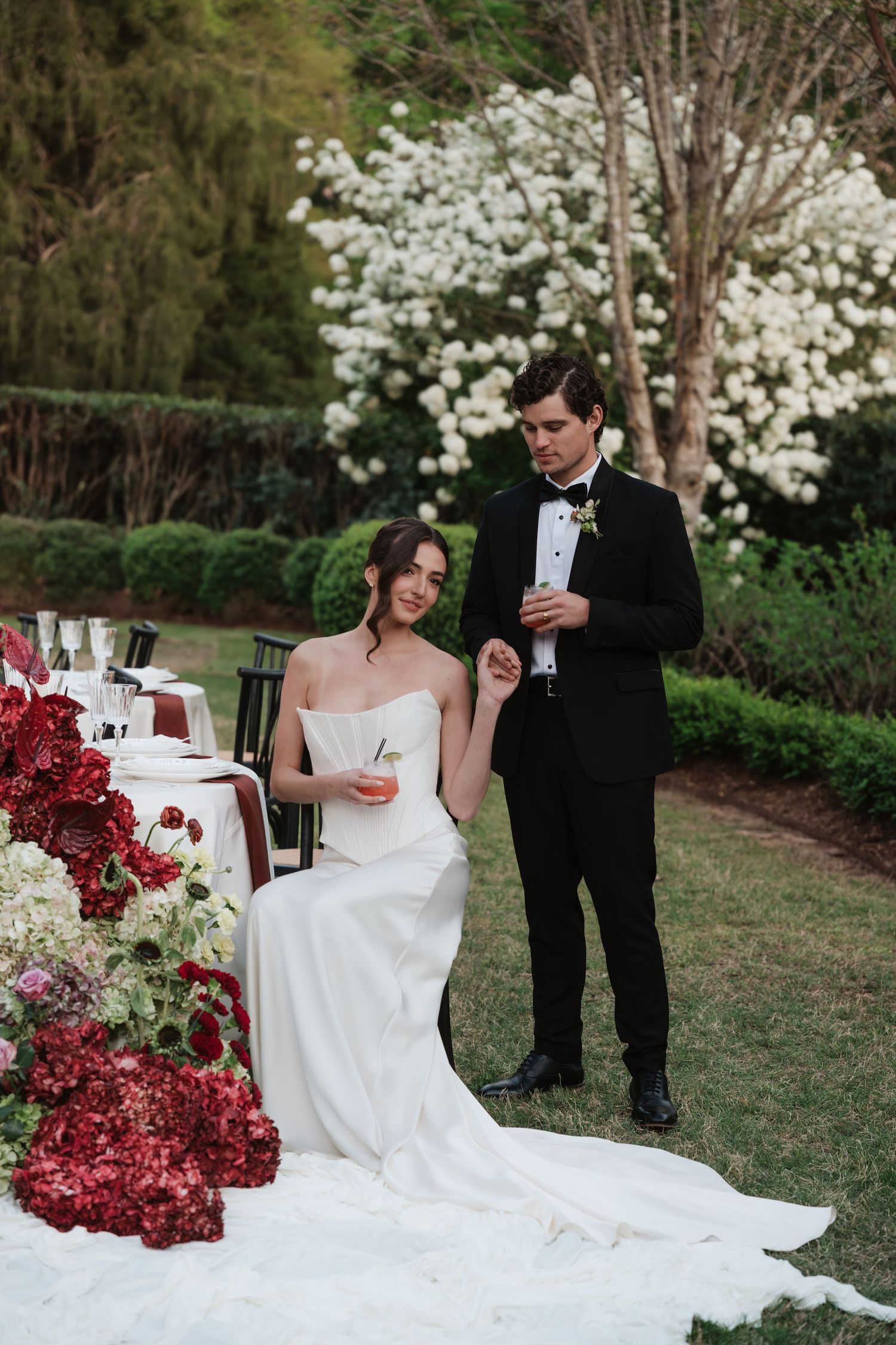 Bride in white gown seated at outdoor wedding table with red florals, groom in black tuxedo beside her.
