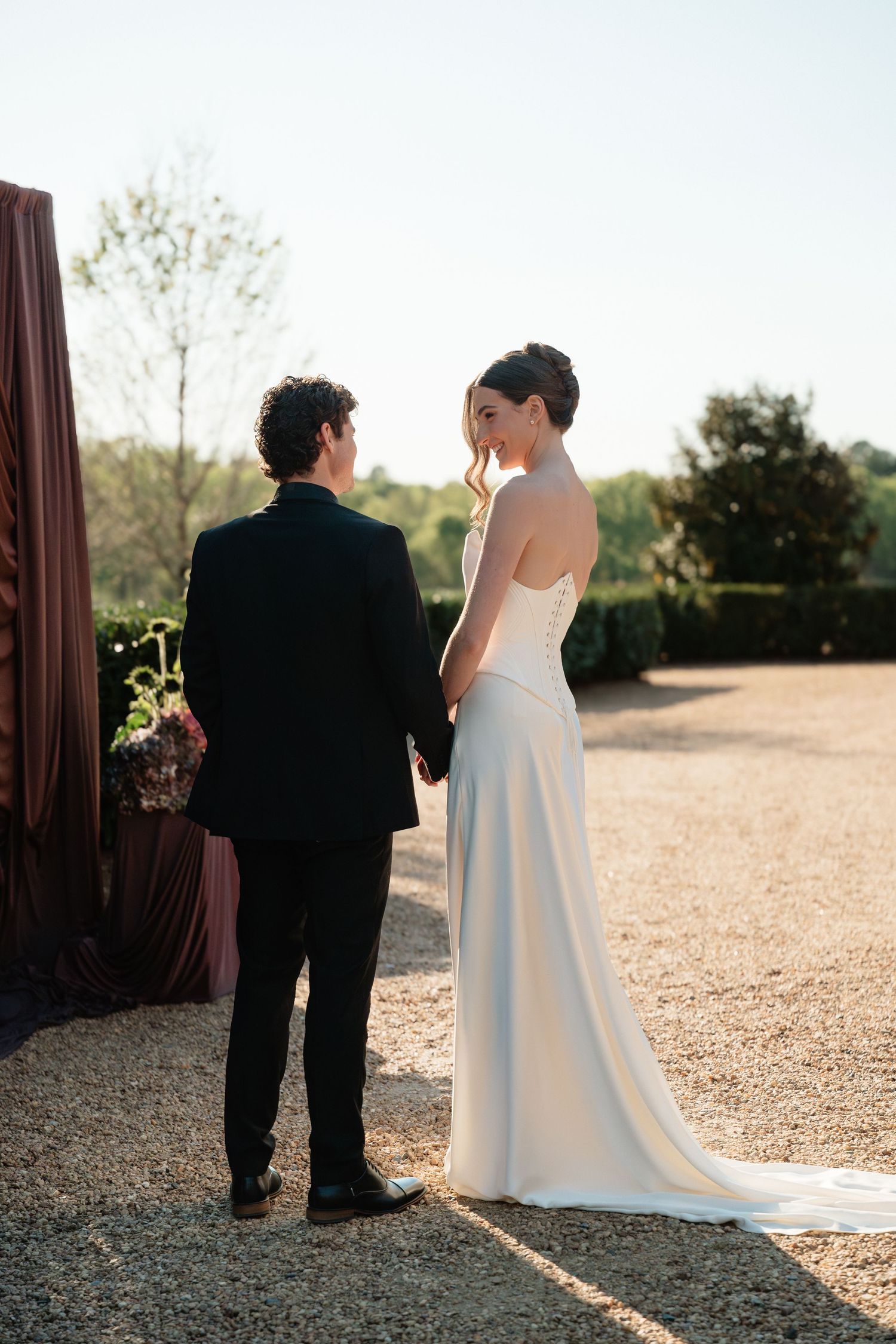 Bride in strapless white gown and groom in black suit hold hands outdoors on gravel path at golden hour wedding.