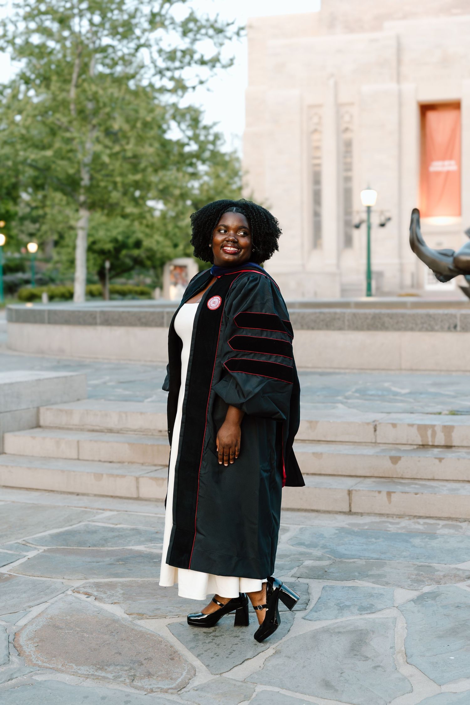 A smiling Black woman in doctoral graduation regalia poses outdoors on a stone plaza near a university building.