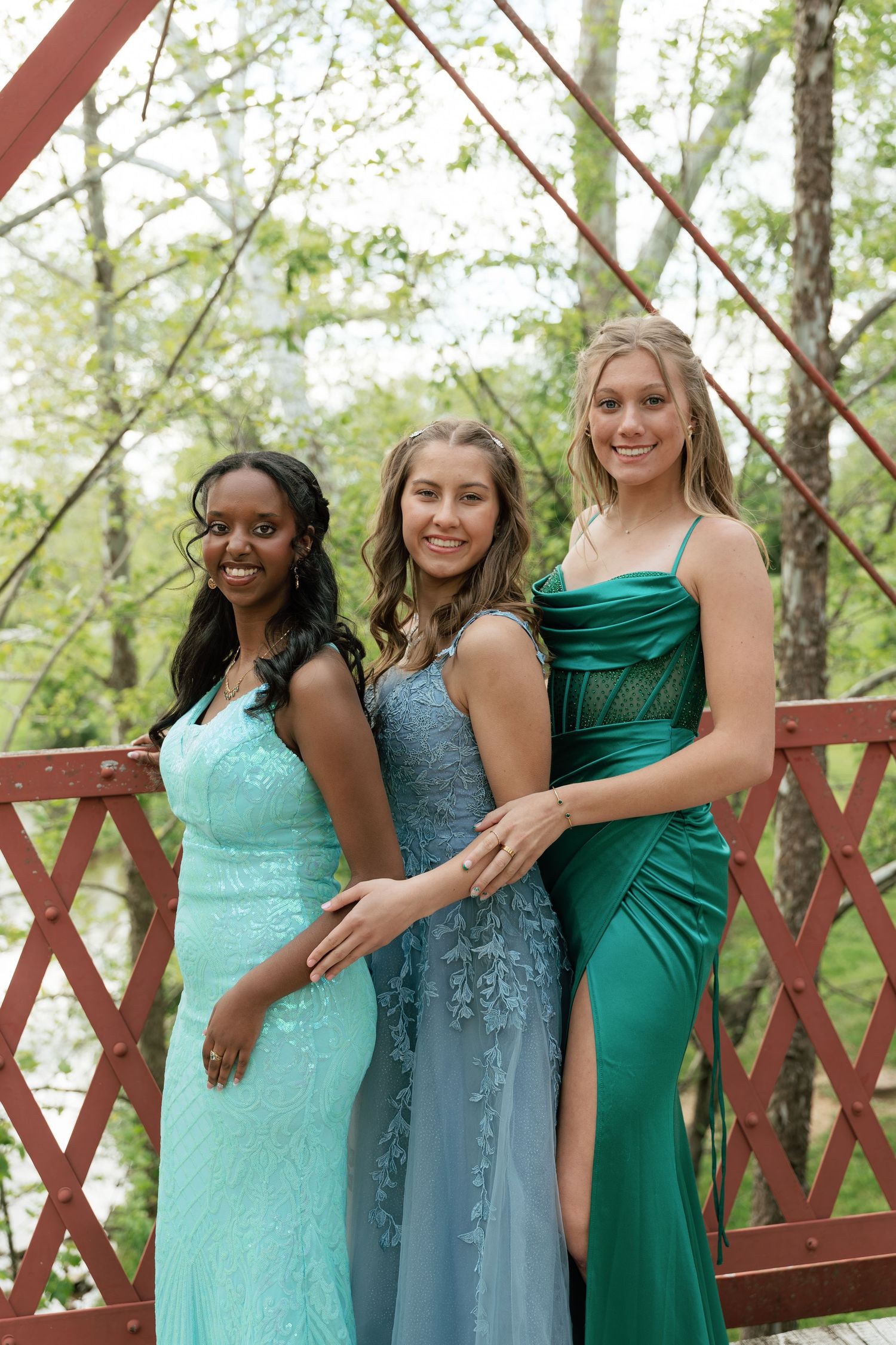 Three teenage girls in elegant prom gowns of mint, blue, and green pose together on a red bridge outdoors.