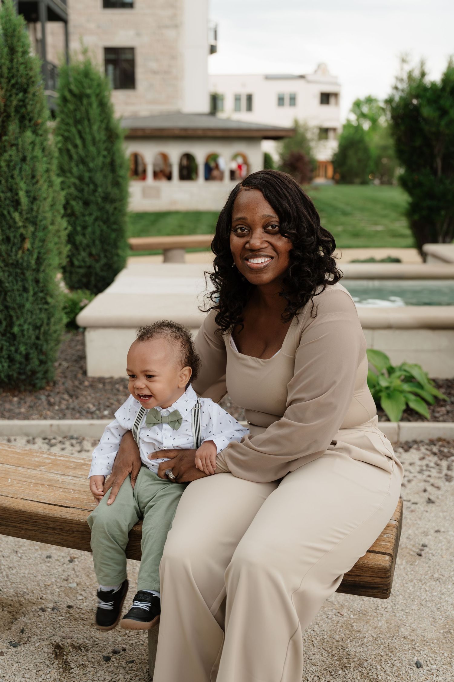 A smiling Black woman in a beige outfit sits on a bench holding a baby boy in a green bow tie suit outdoors.