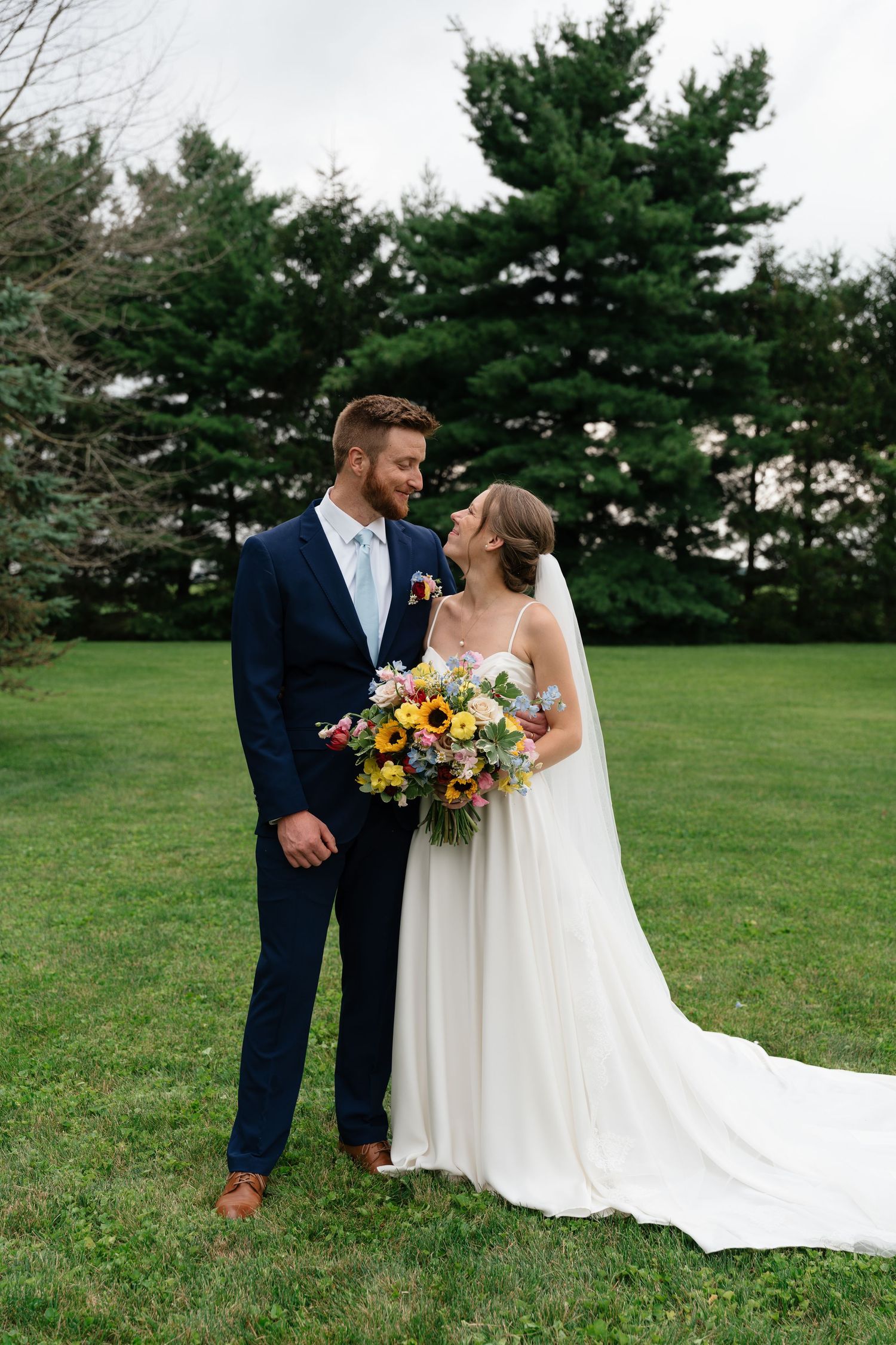 Bride and groom share a romantic moment on green lawn, she holds colorful sunflower bouquet, tall evergreen trees behind them.