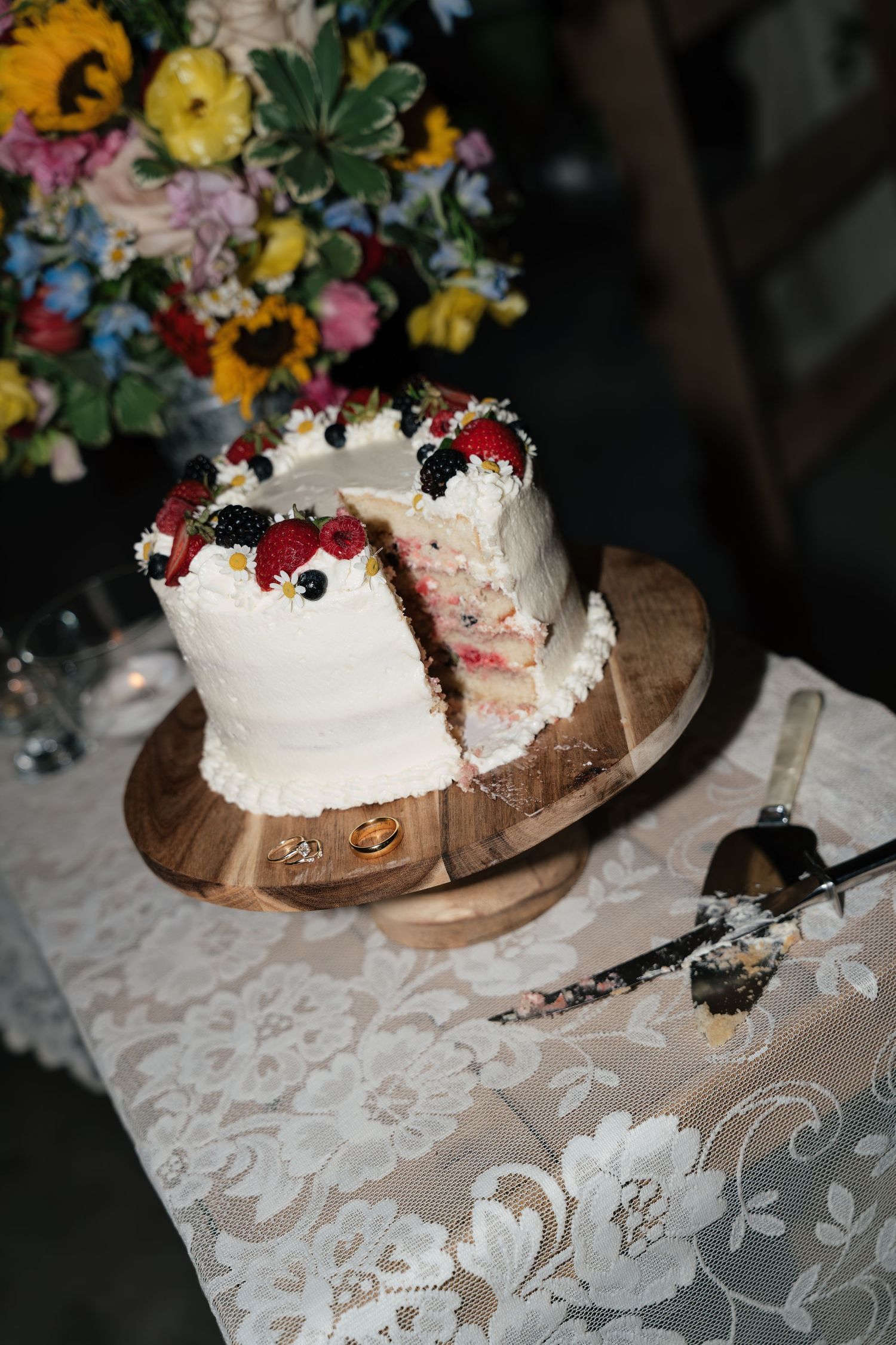 White wedding cake with fresh berries on wooden stand, slice removed, beside cake server on lace tablecloth.