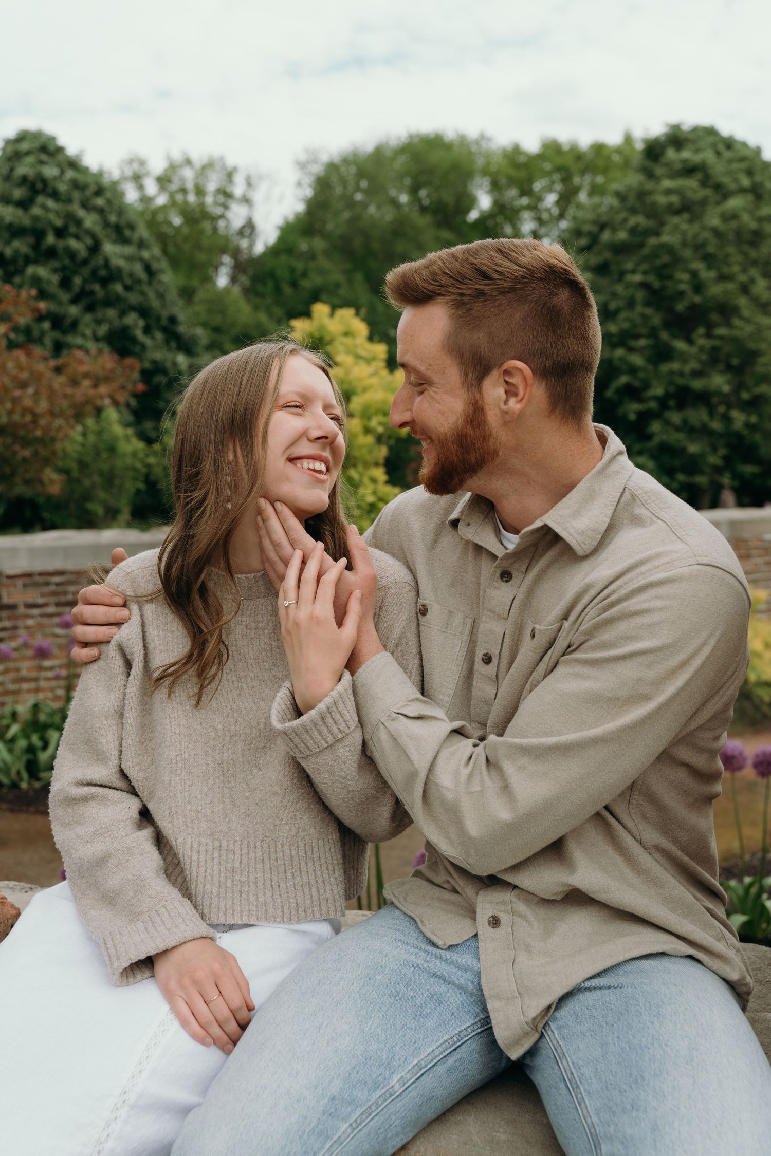A smiling couple sits outdoors in a garden, sharing a joyful moment as the man gently touches the woman's face.