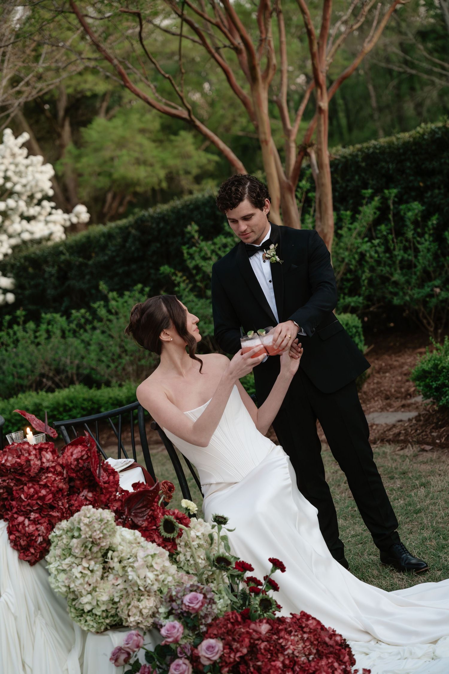 Bride in white gown and groom in black tuxedo share a toast amid lush garden florals of red and white blooms.