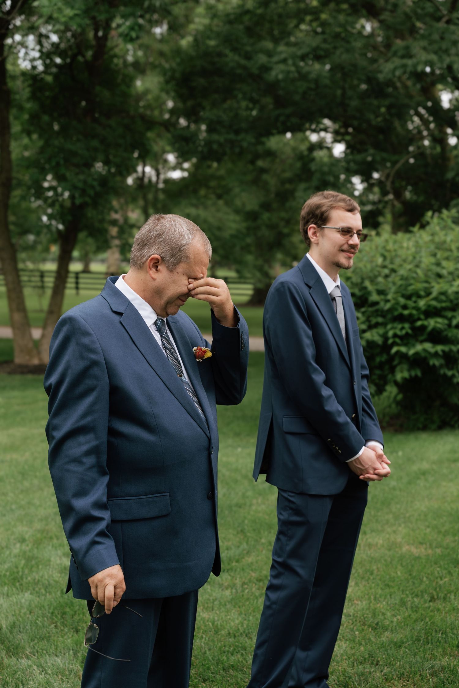 Two men in navy suits at a wedding, one emotional wiping tears while the other smiles, standing on green grass.