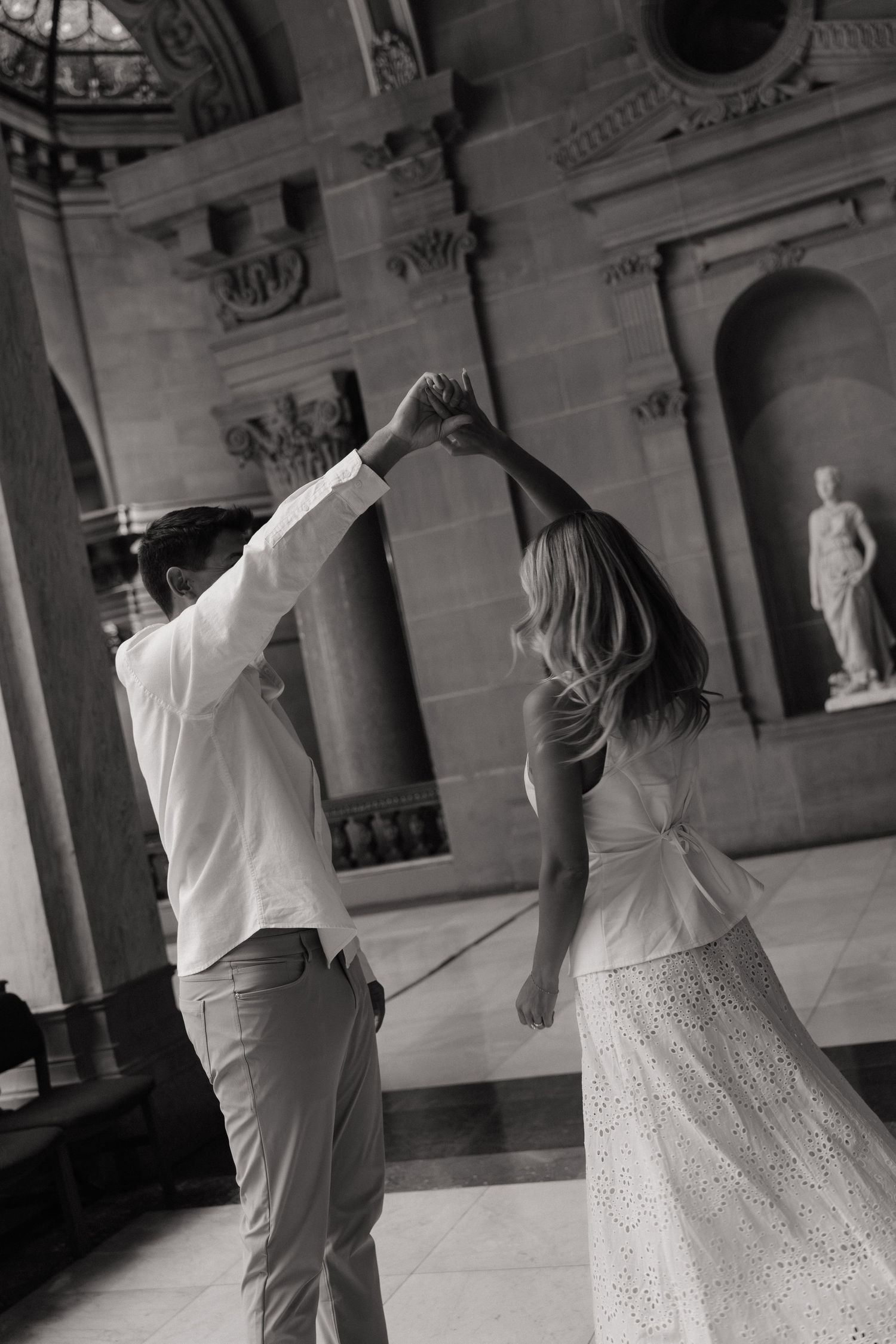 Black and white photo of a couple dancing elegantly in a grand classical building with marble statues.