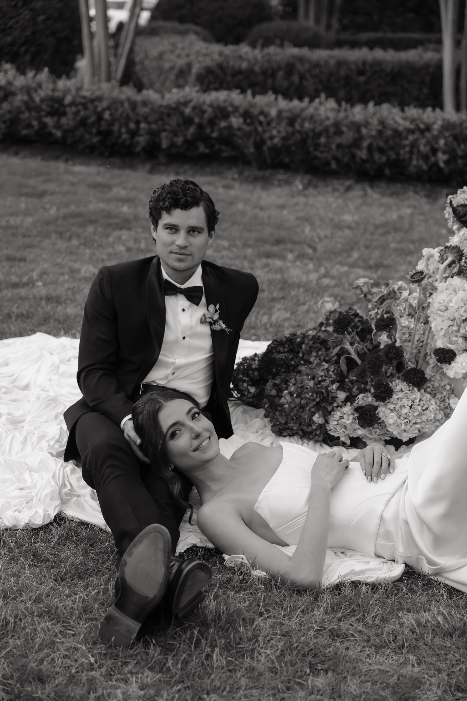 Black and white photo of bride and groom sitting on grass, groom in tuxedo, bride in wedding gown with floral bouquet.