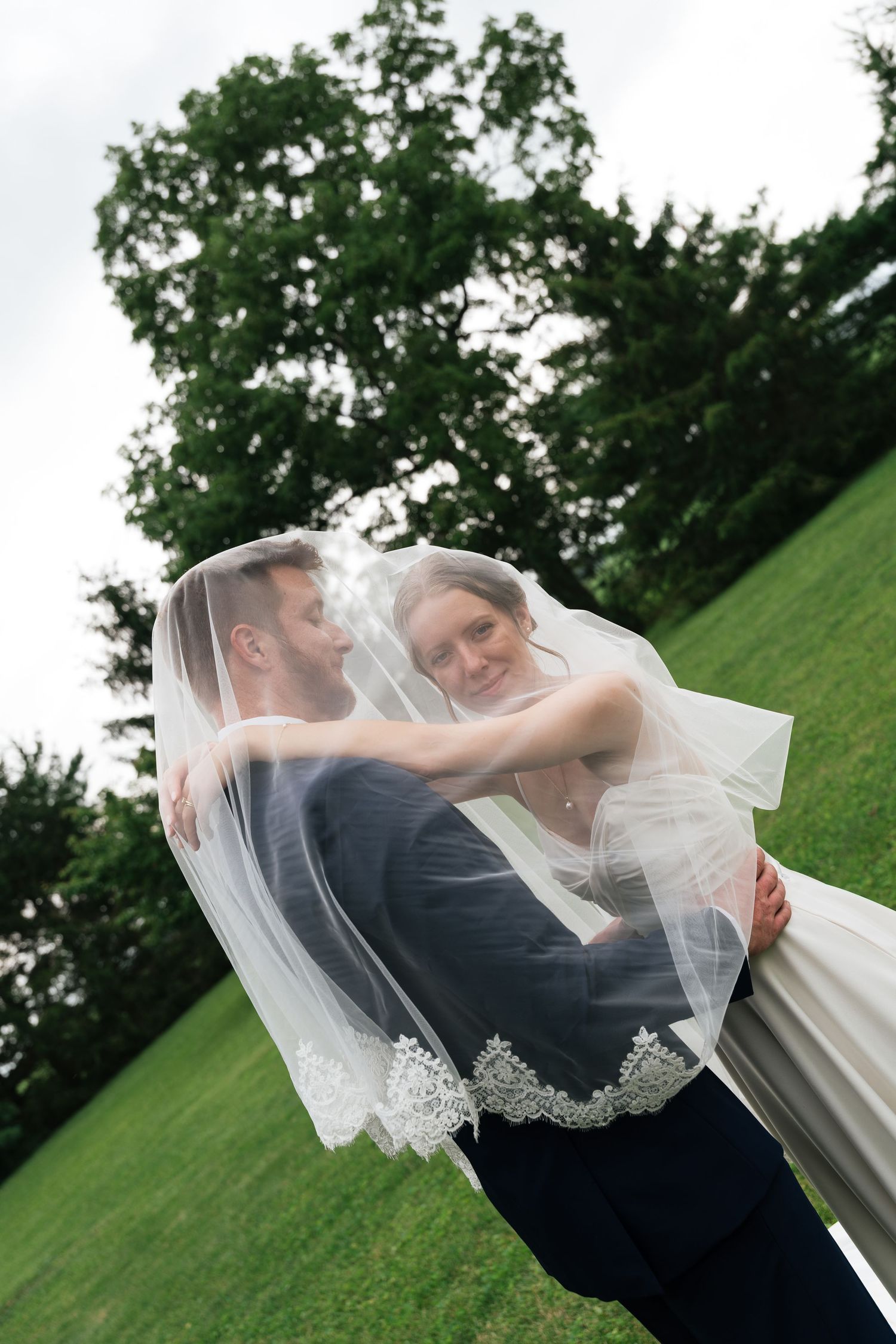 Bride and groom share romantic moment under lace wedding veil outdoors on green lawn with trees.