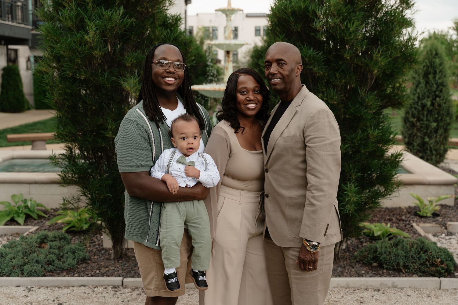Family of four posing outdoors in coordinated neutral outfits, with a man holding a baby, in a lush garden setting.