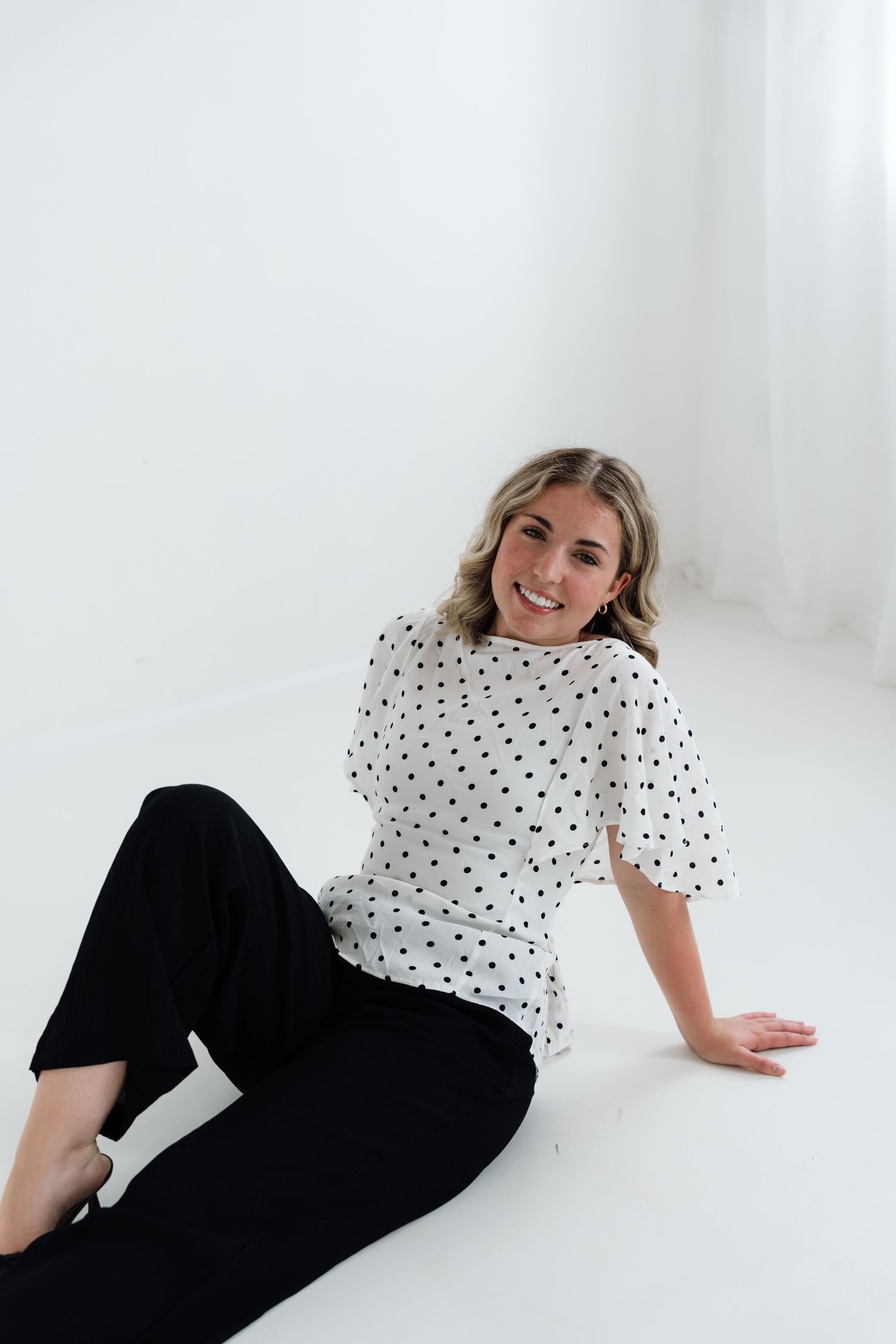 Woman in white polka dot top and black pants sitting on white floor, smiling at camera.
