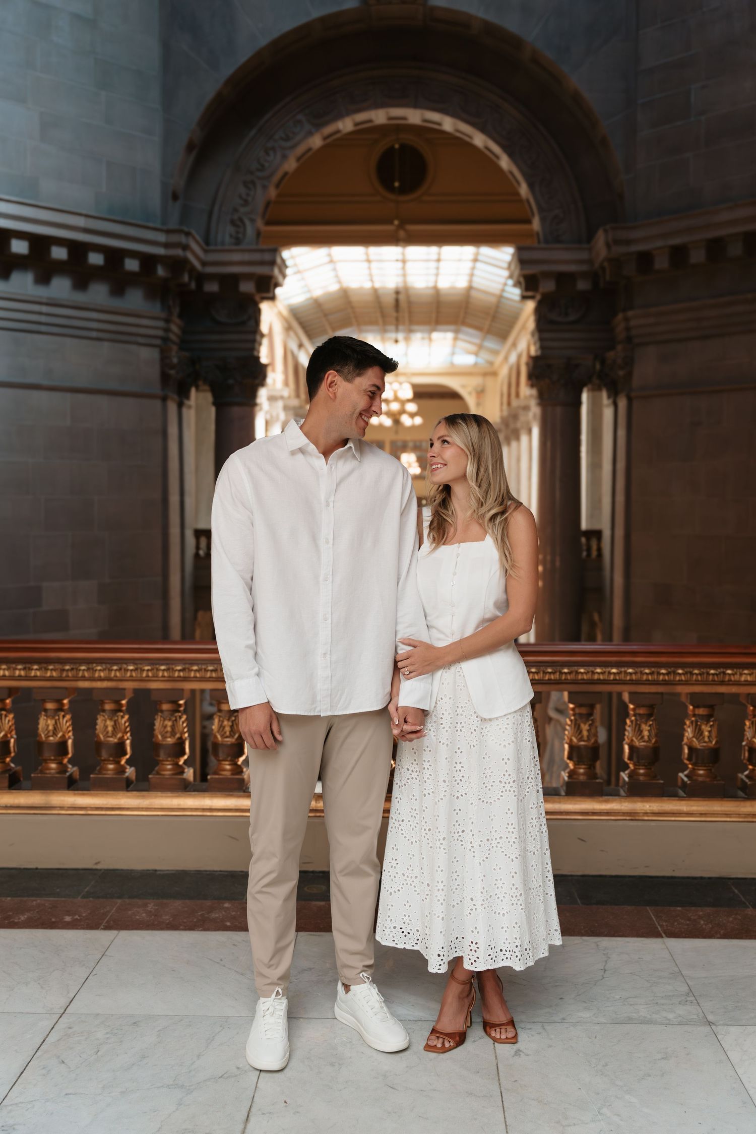 A couple in white outfits smiling at each other inside an elegant neoclassical building with arched ceilings.