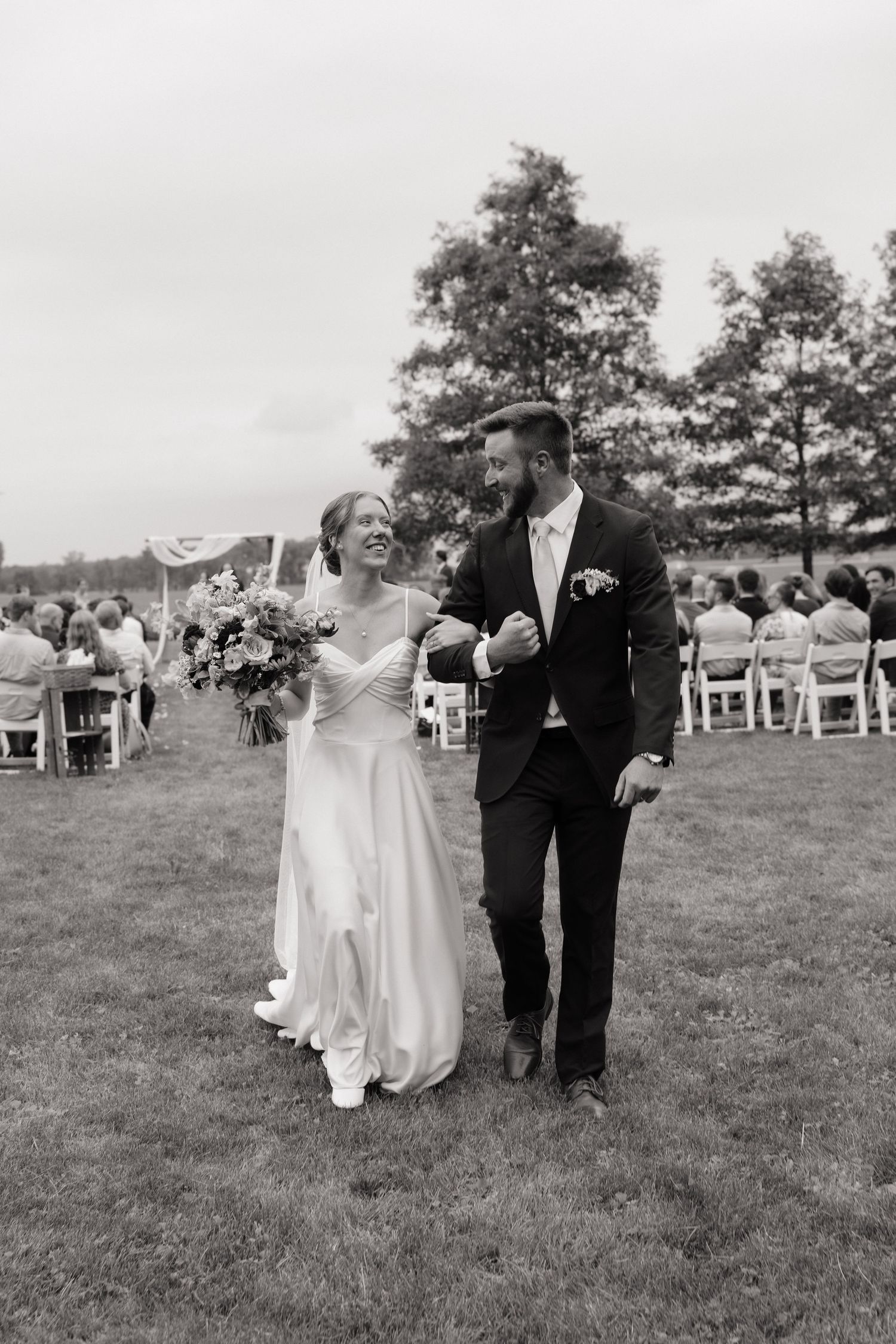 Black and white photo of a smiling bride and groom walking down the aisle after their outdoor wedding ceremony.
