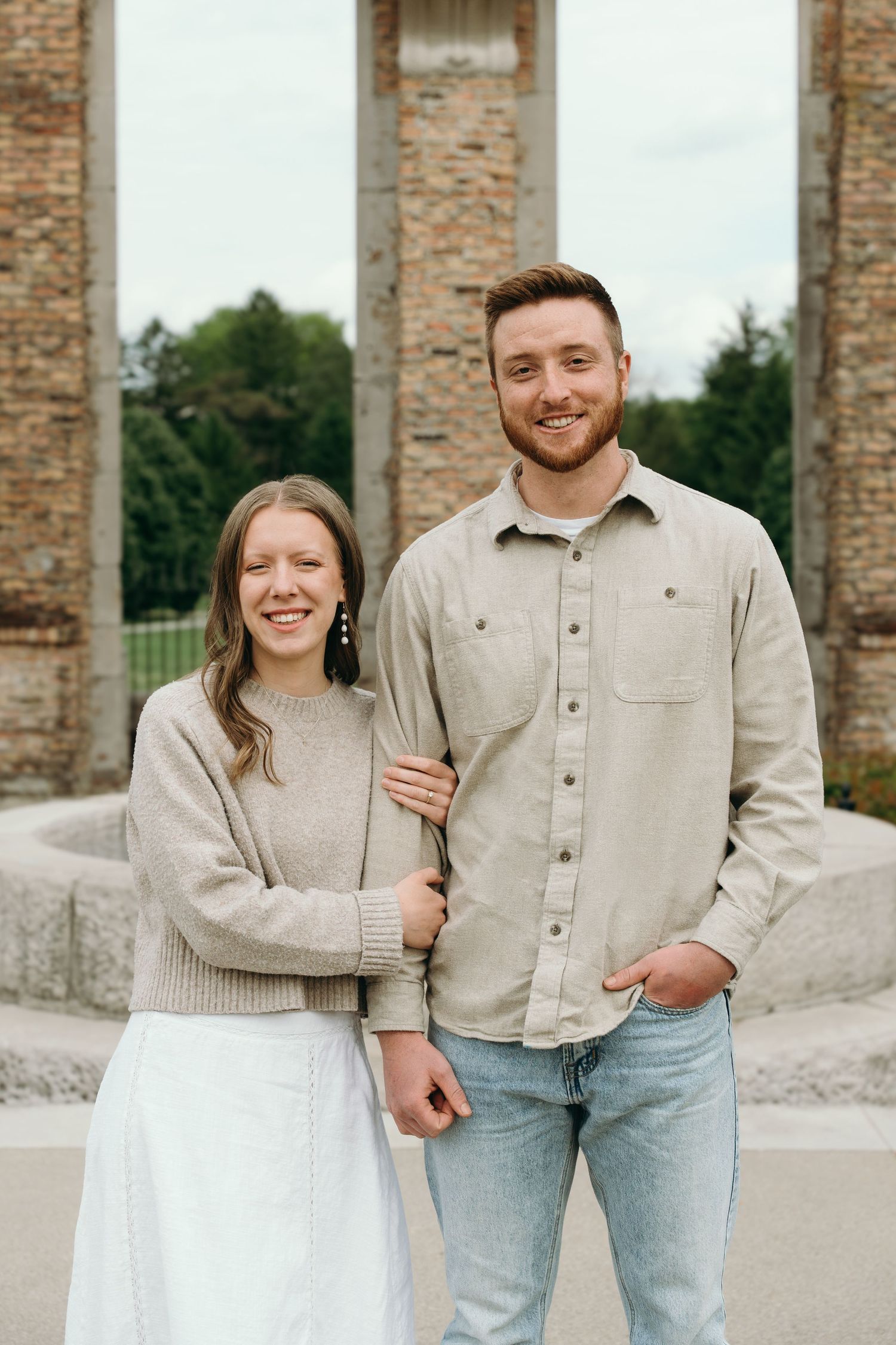 Smiling couple posing outdoors near stone columns, woman in beige sweater and white skirt, man in khaki shirt and jeans.