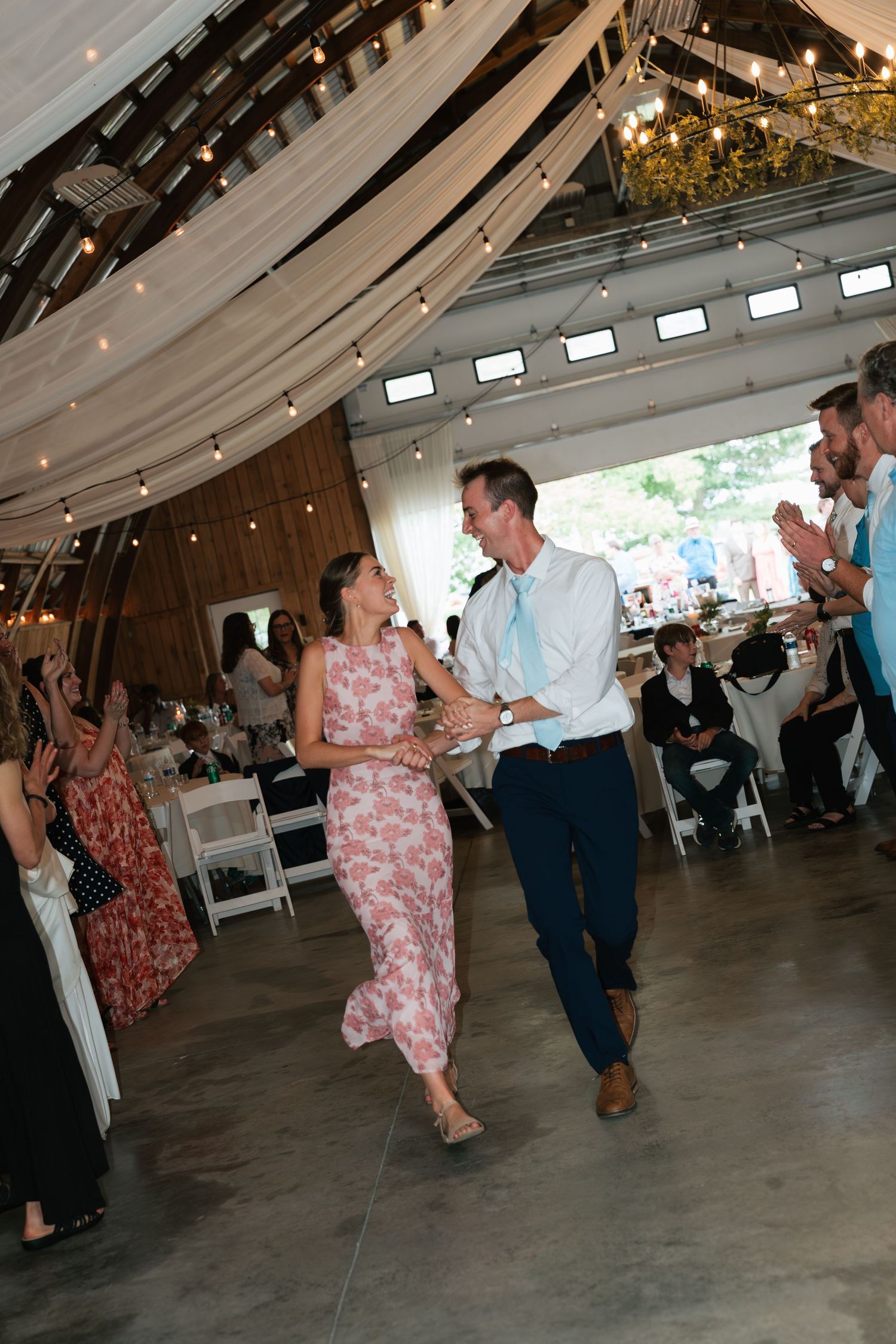 Couple dancing joyfully at a wedding reception in a decorated barn venue with string lights and draped fabric.