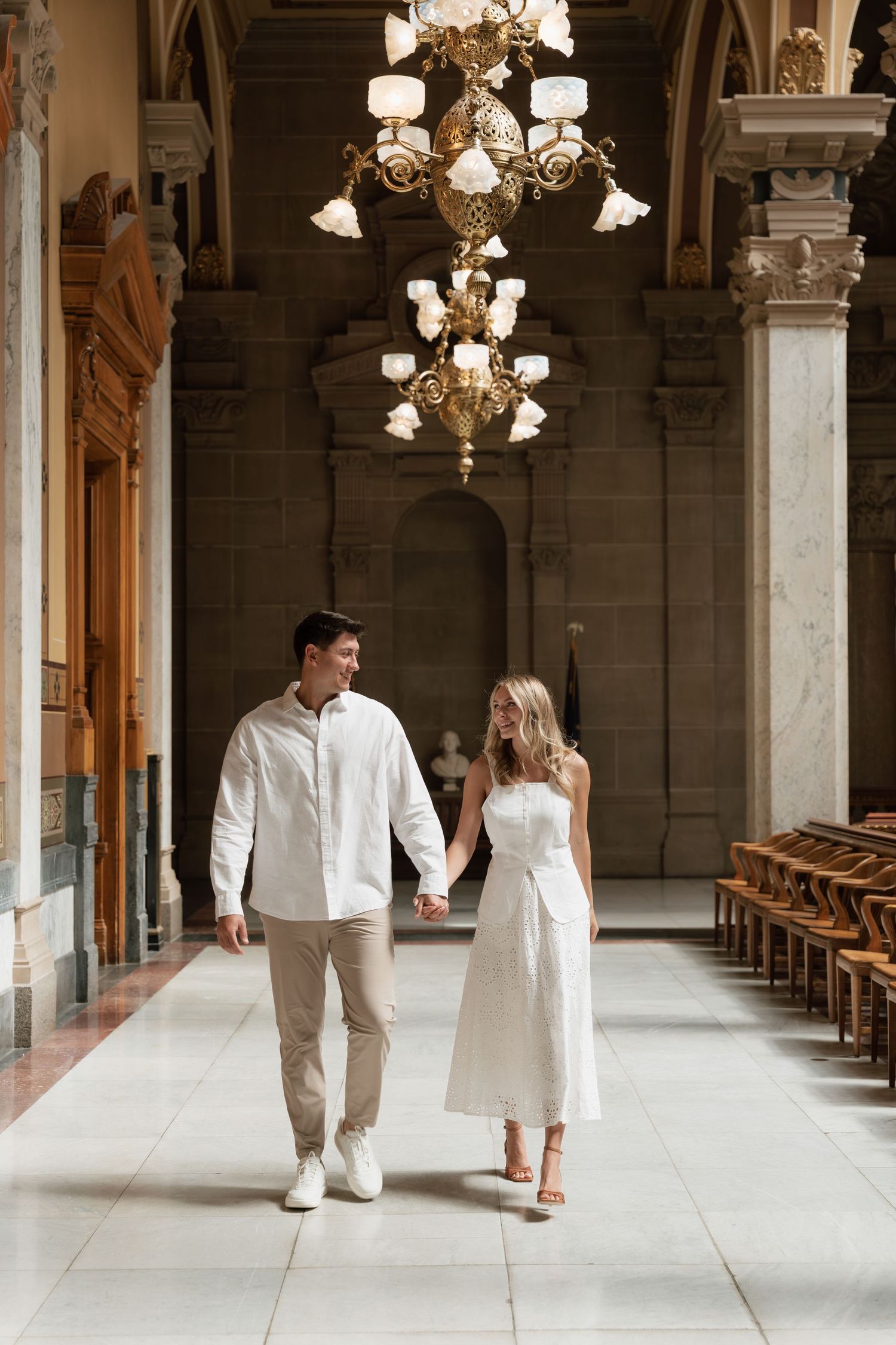 Couple holding hands walking through elegant hall with ornate chandelier and marble columns in white outfits.