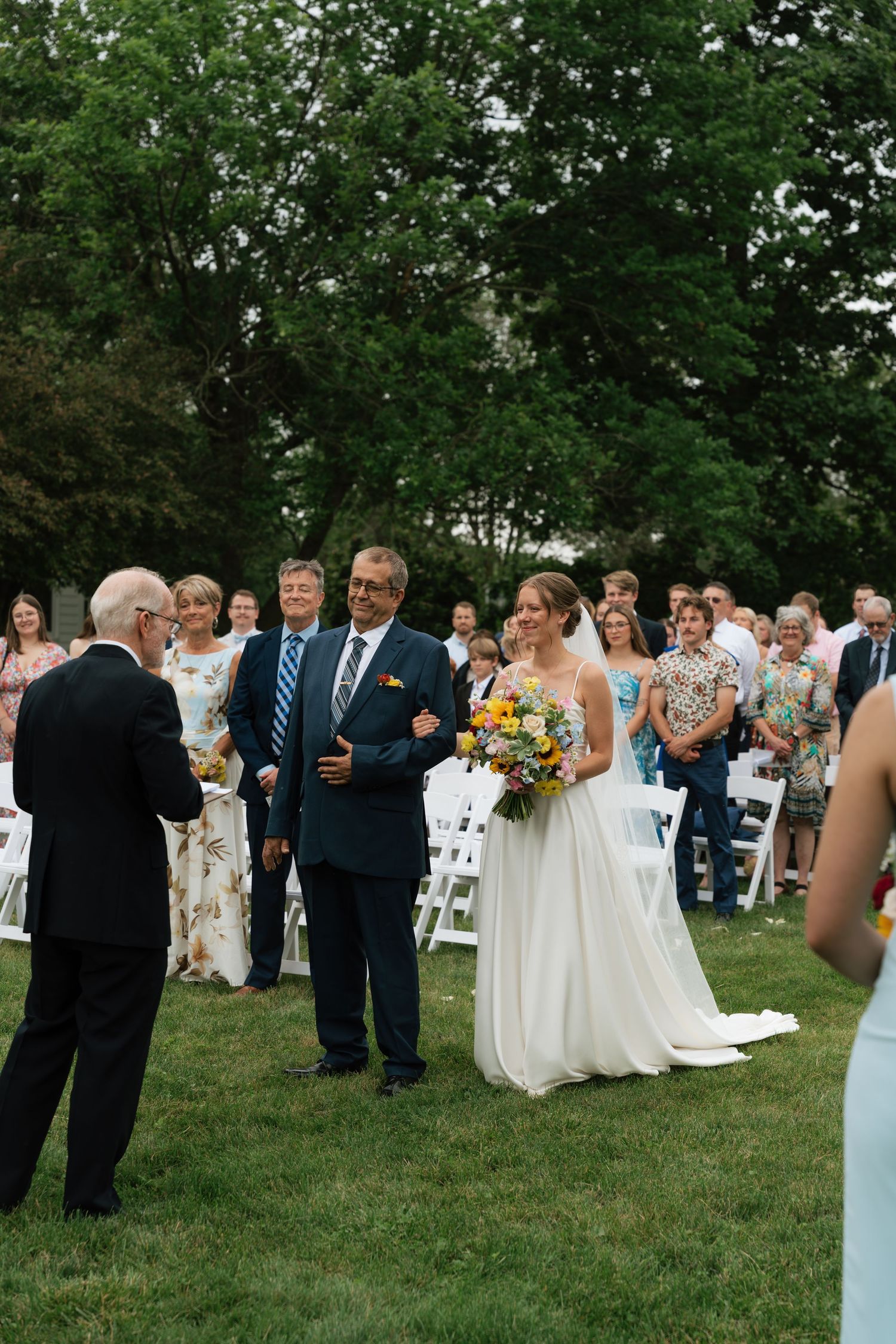 Bride walks down the aisle with her father at an outdoor garden wedding ceremony with seated guests.