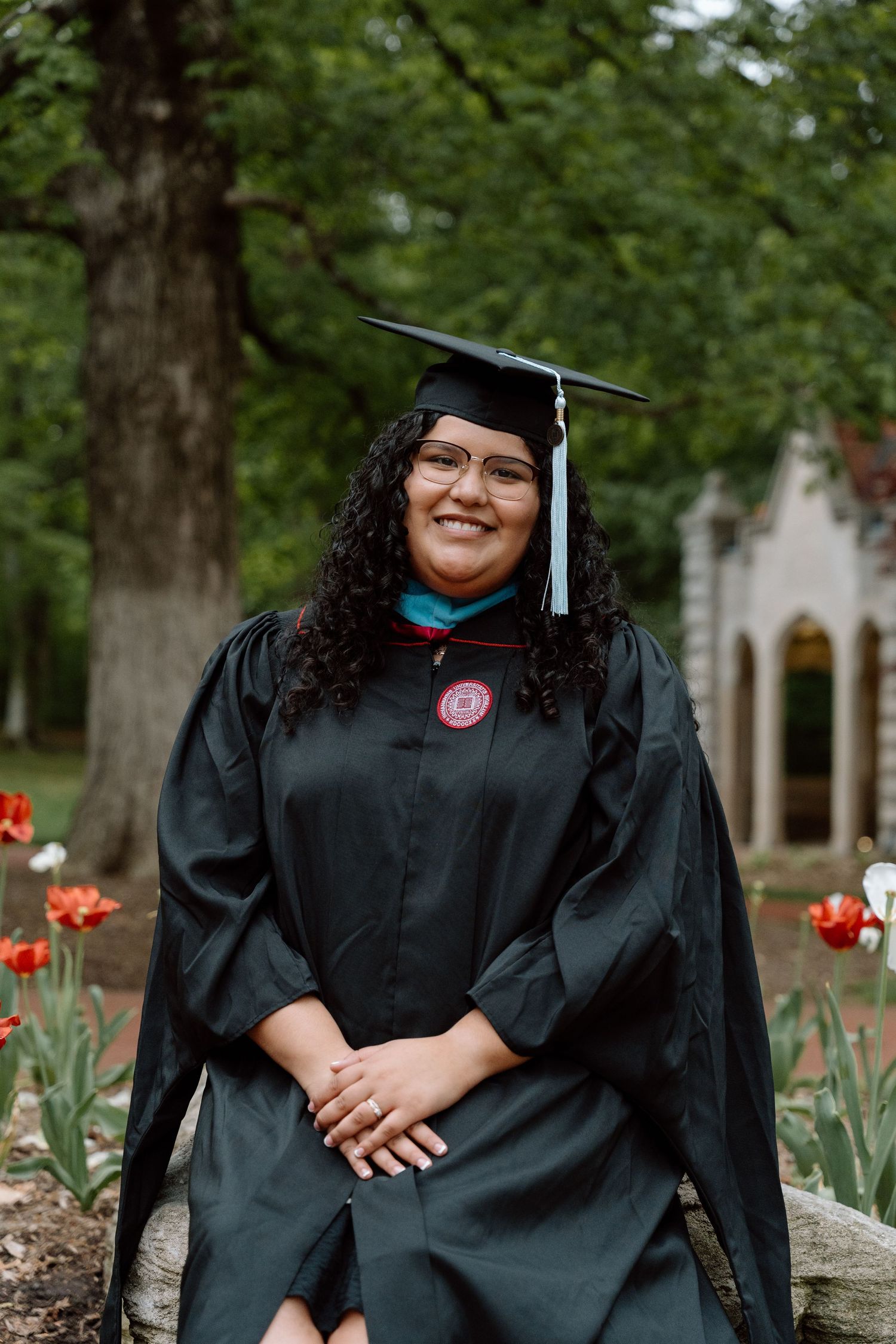 Smiling graduate in black cap and gown poses outdoors among red tulips on a college campus.