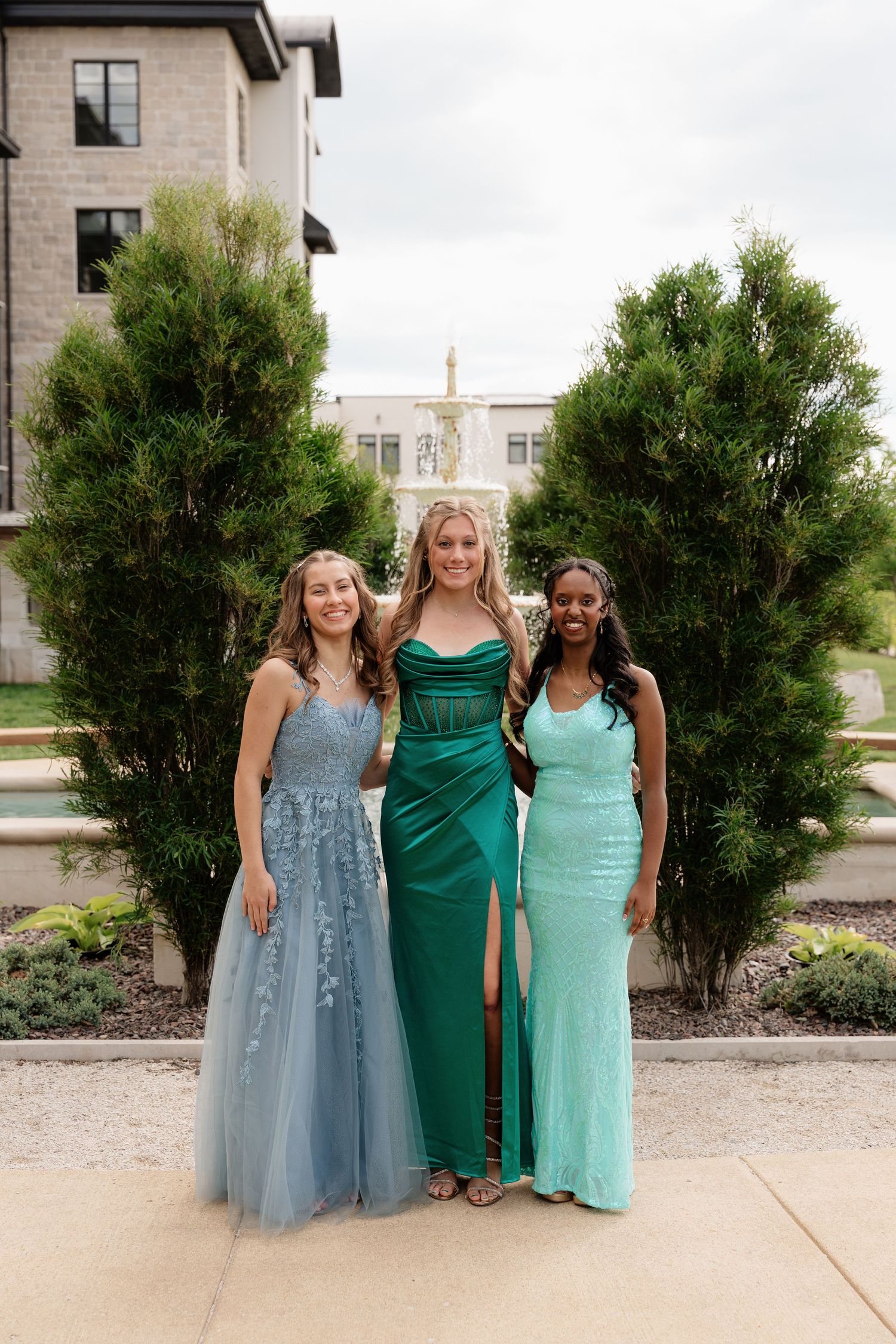 Three young women in elegant prom gowns posing outdoors near a fountain.
