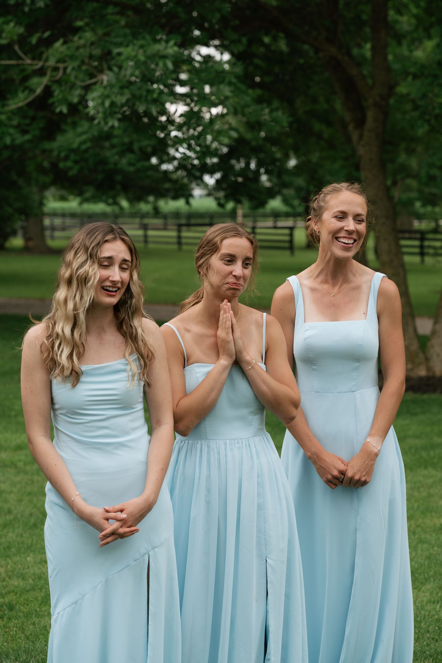 Three bridesmaids in matching light blue dresses smile and laugh together at an outdoor wedding.