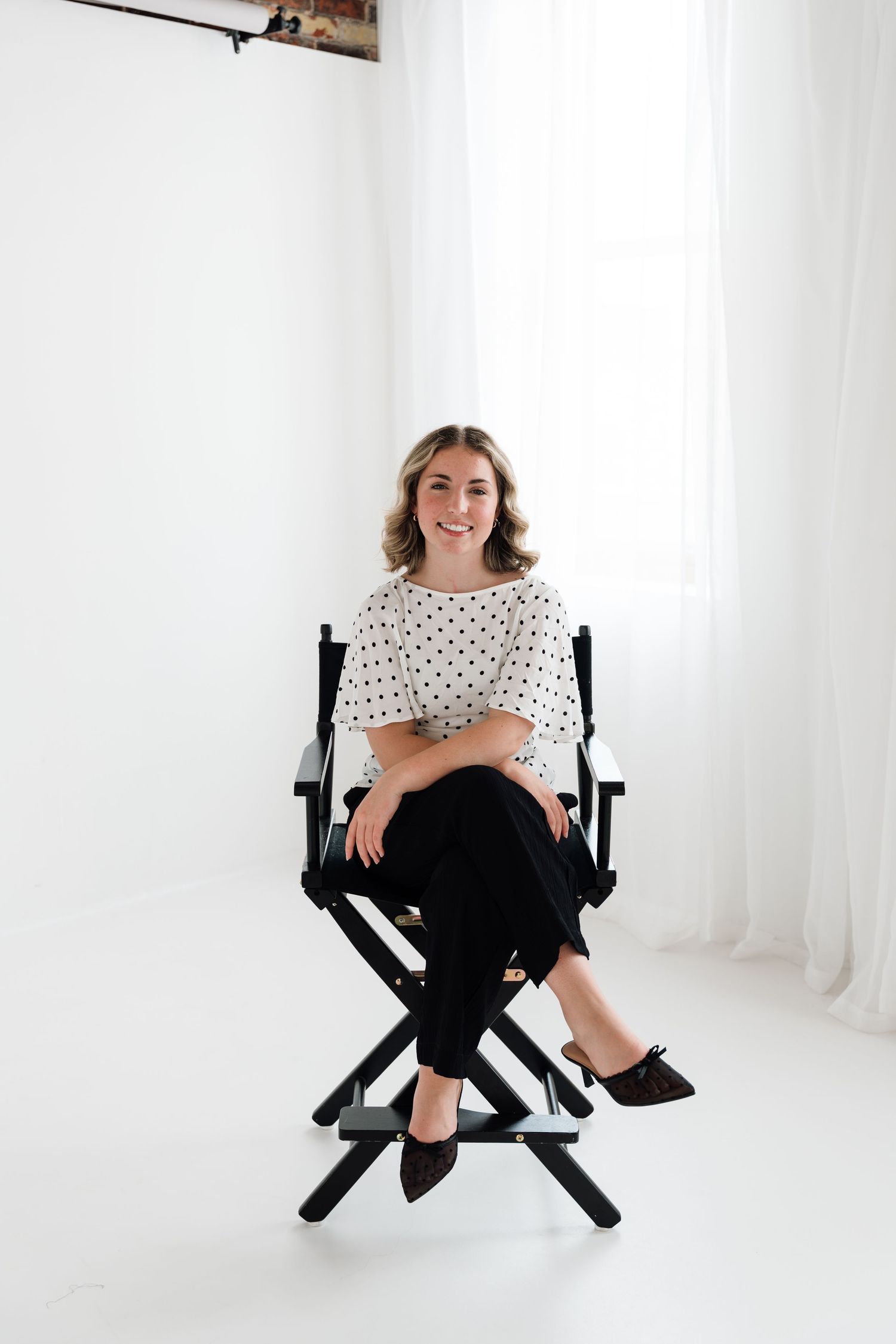 Smiling woman in polka dot blouse and black pants sitting in a director's chair in a white studio.
