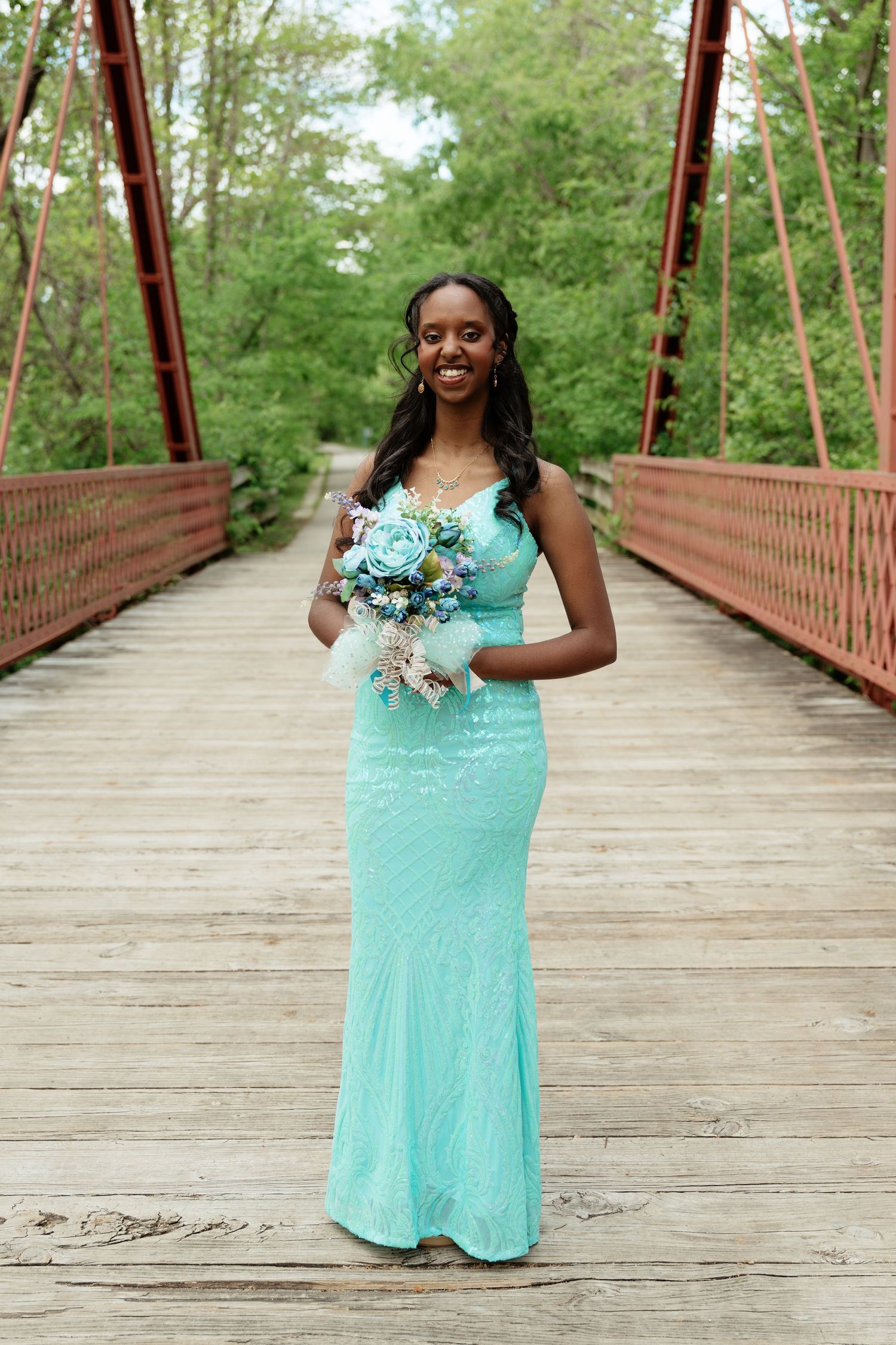 Young woman in a teal sequin gown holding a blue bouquet on a wooden bridge.