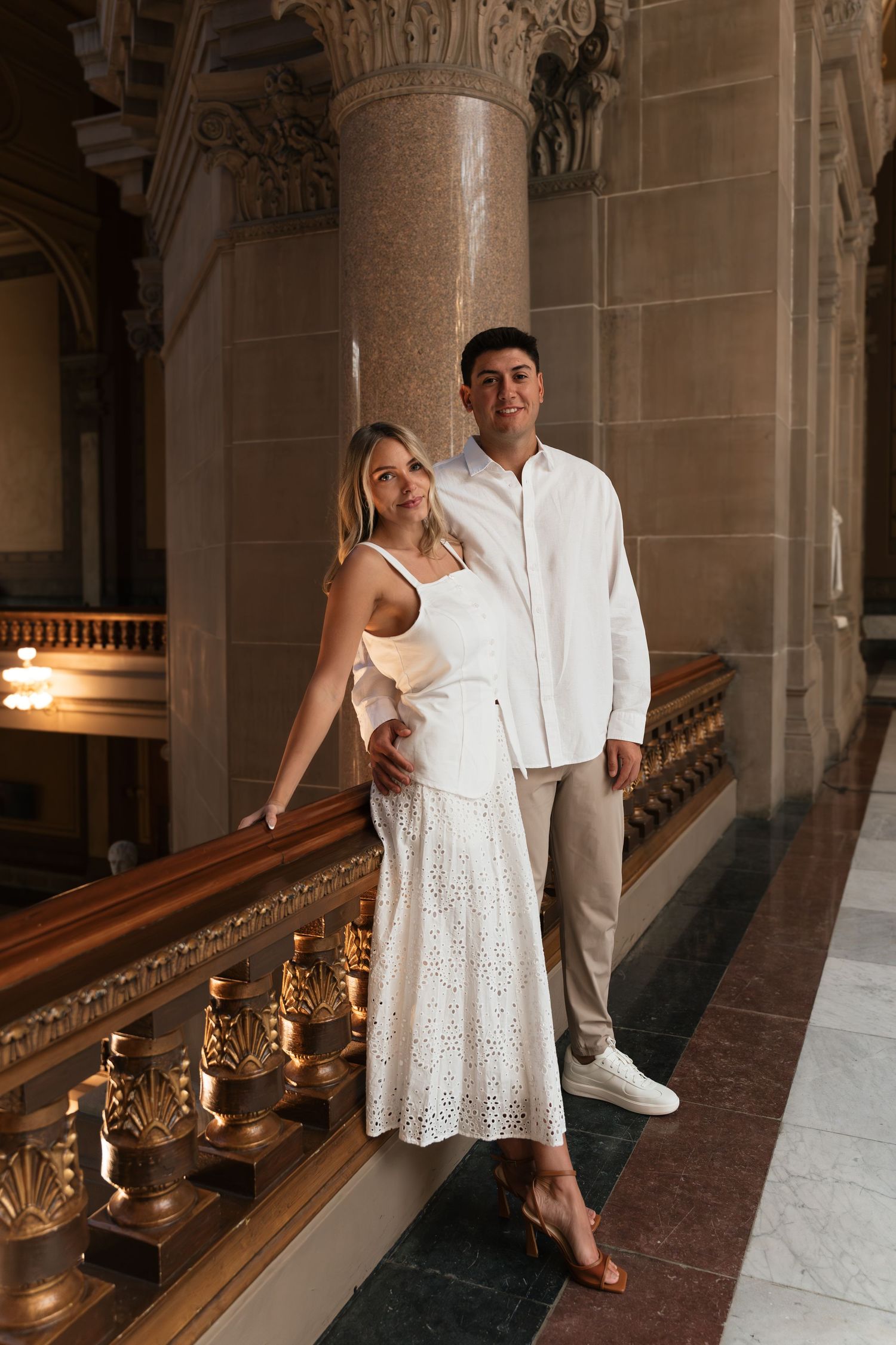 Elegant couple posing in white outfits beside ornate golden railings in a grand architectural interior.