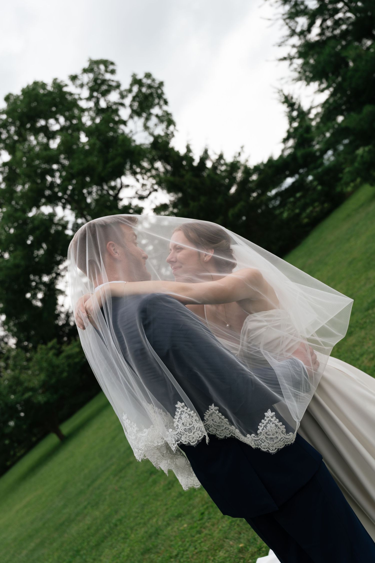 Groom lifts bride under flowing lace veil on green lawn, sharing romantic moment at outdoor wedding ceremony.