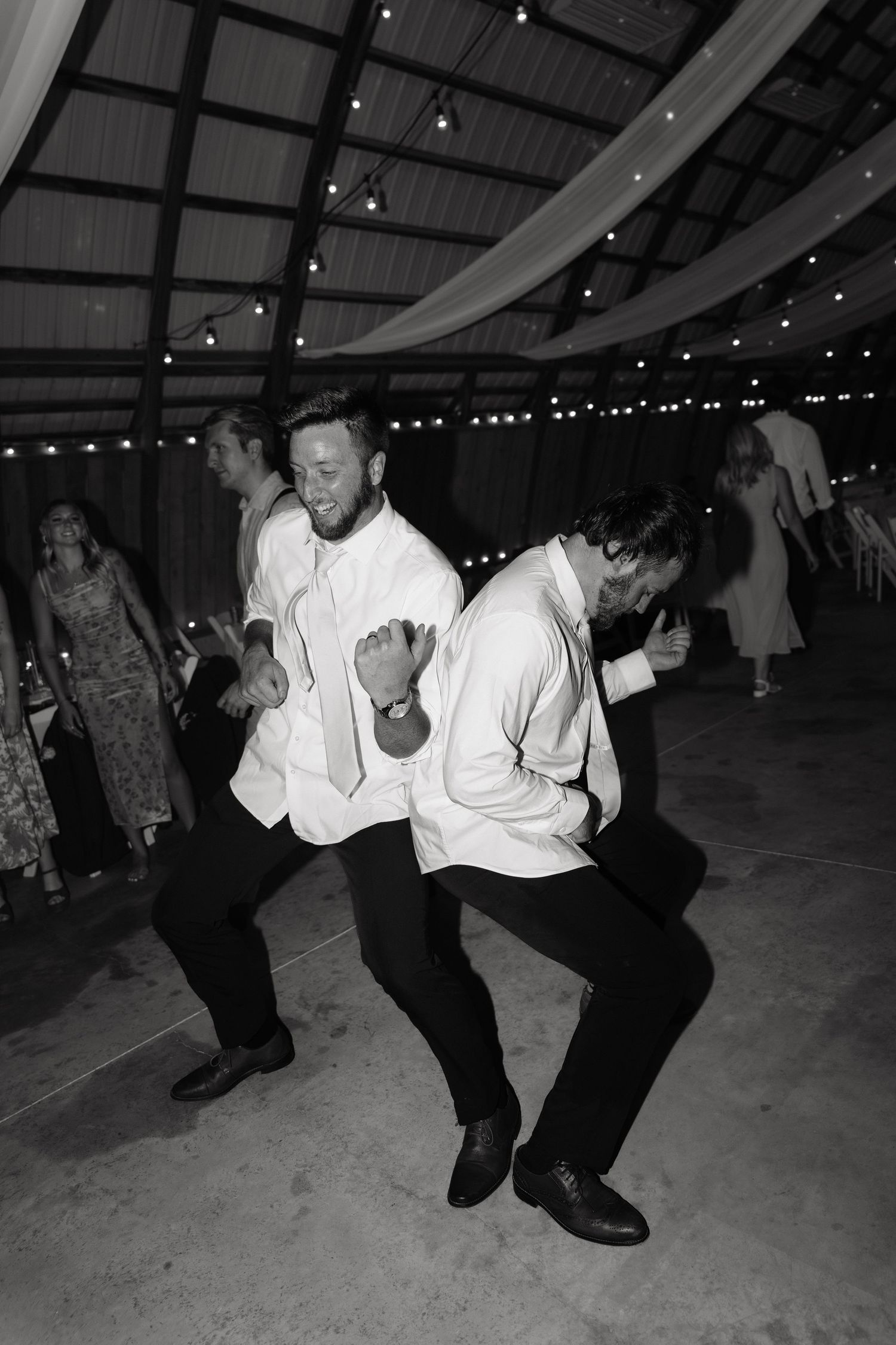 Two groomsmen dance energetically at a wedding reception in a decorated barn venue, captured in black and white.