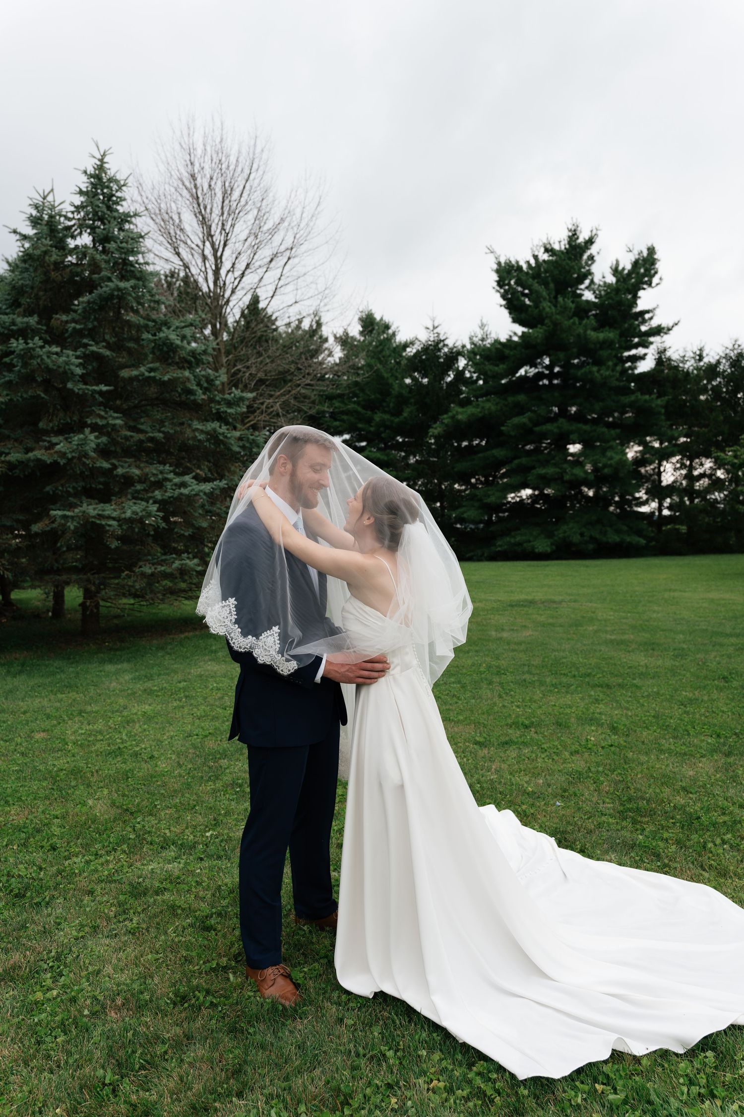 Bride and groom sharing a romantic moment under a wedding veil on a green lawn surrounded by evergreen trees.