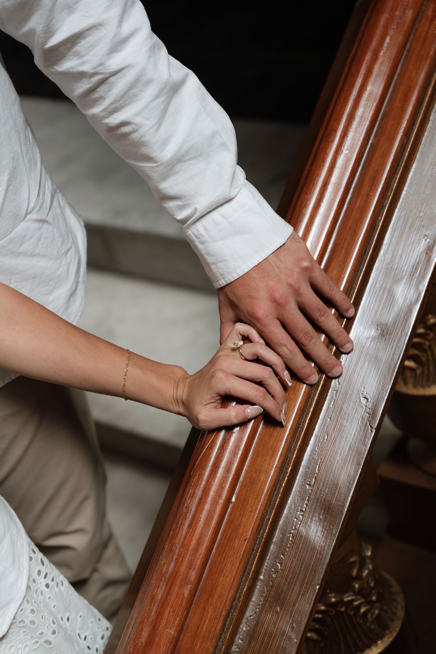 Couple holding hands on a wooden stair railing, wearing white outfits and a delicate bracelet, symbolizing love and togetherness.