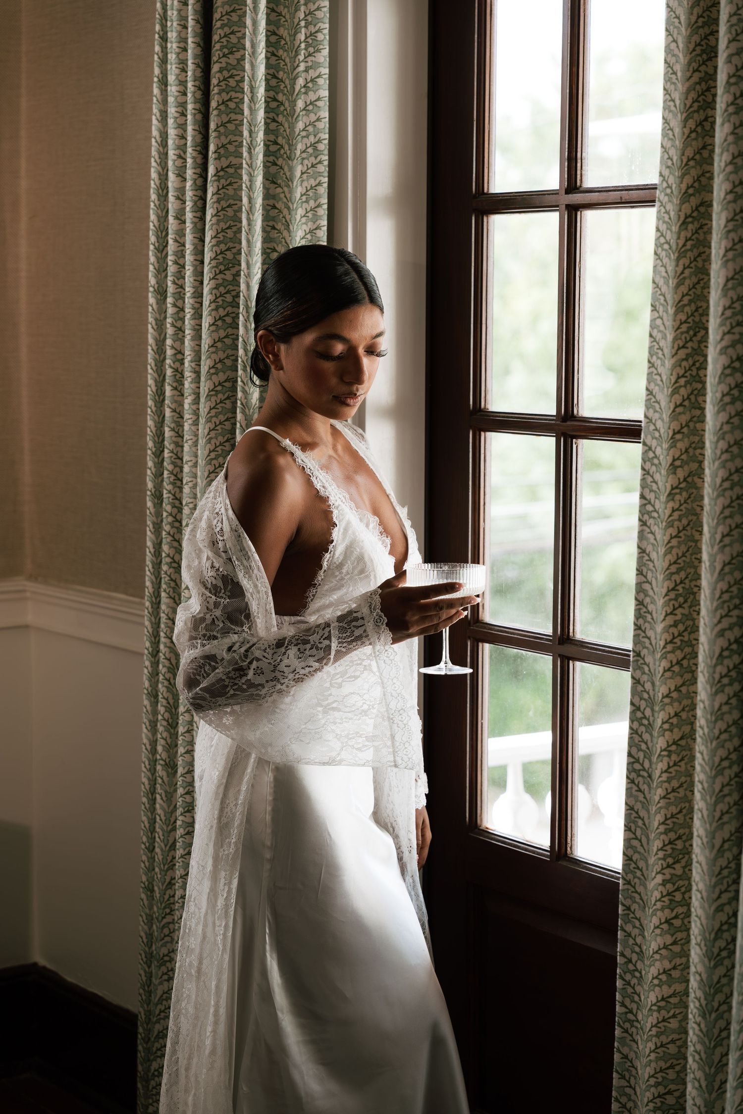 Bride in white lace wedding gown holding a cocktail glass by a sunlit window with green curtains.