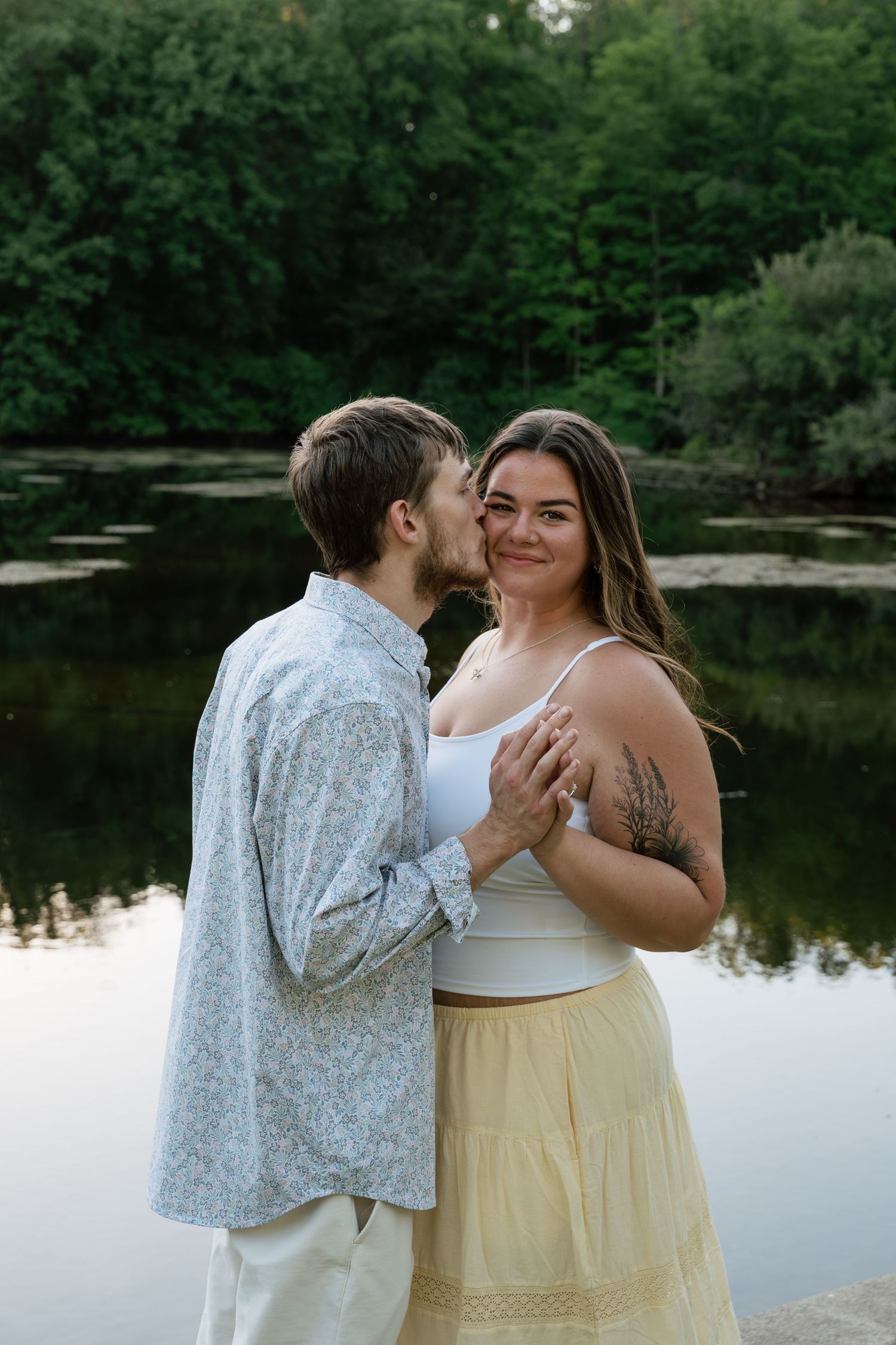 Couple embracing by a serene lake surrounded by lush green trees, woman smiling in white top and yellow skirt.