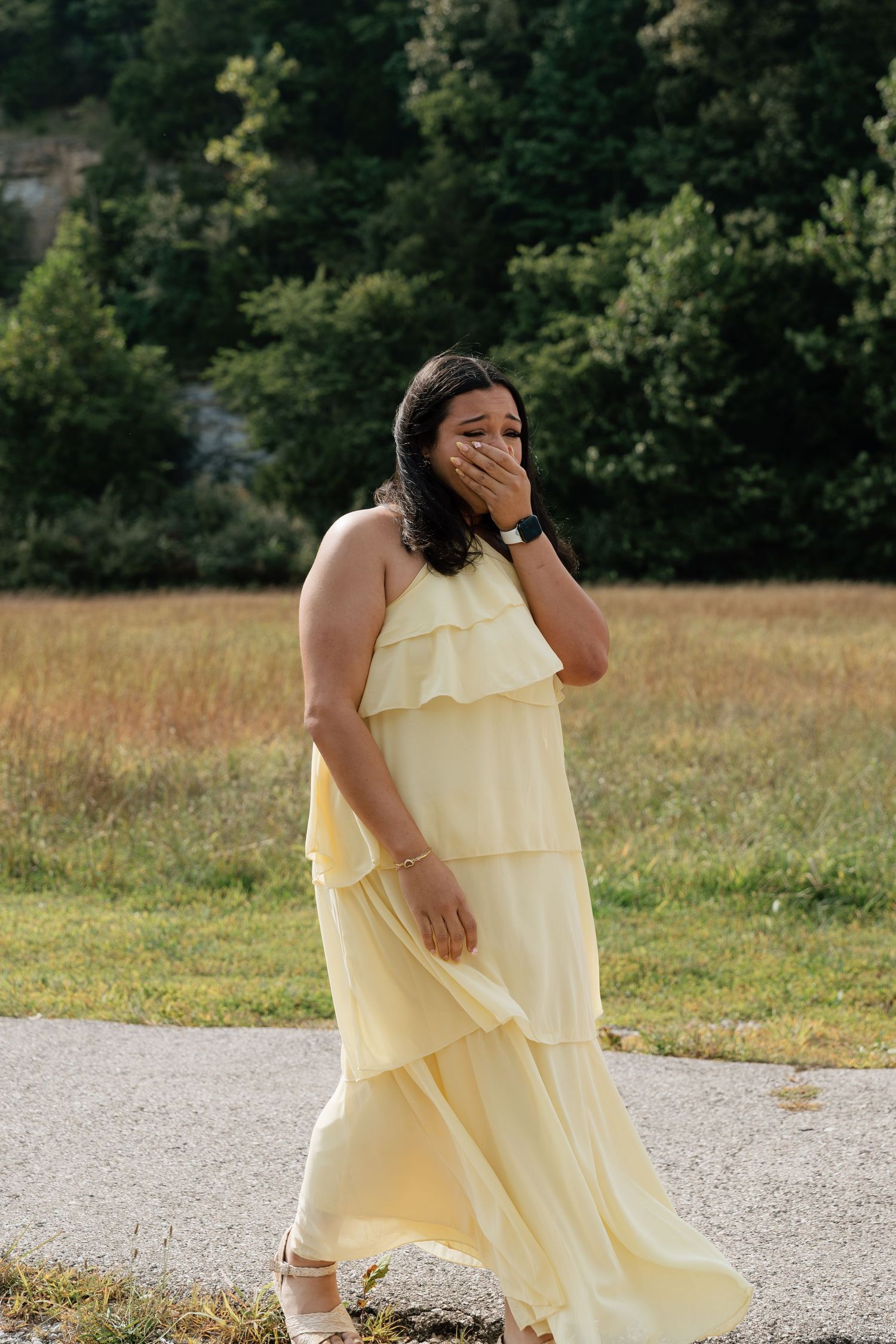 Woman in yellow ruffled maxi dress covering her mouth while walking outdoors near a grassy field.