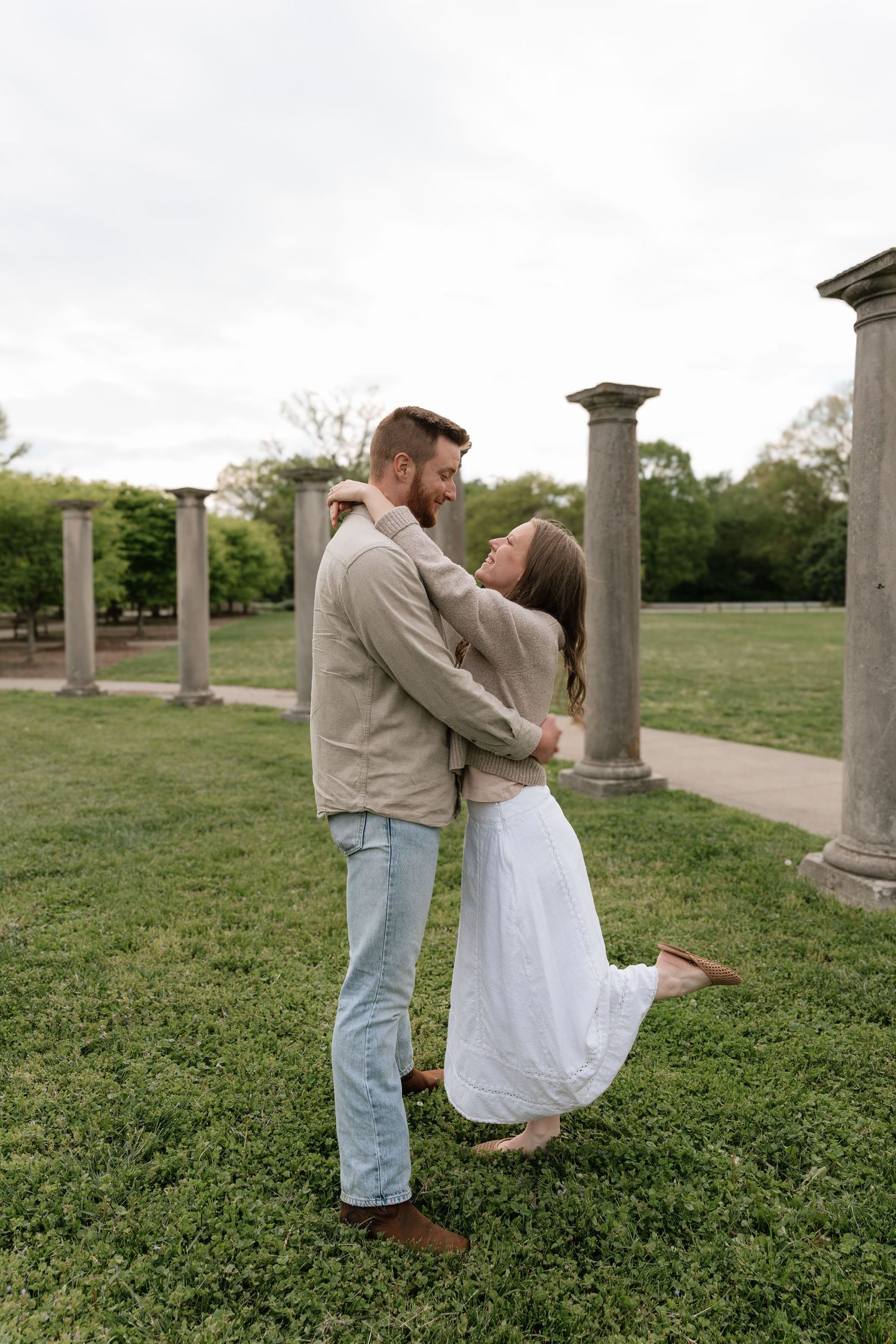 Joyful couple dancing and embracing on green grass among ancient stone columns outdoors.