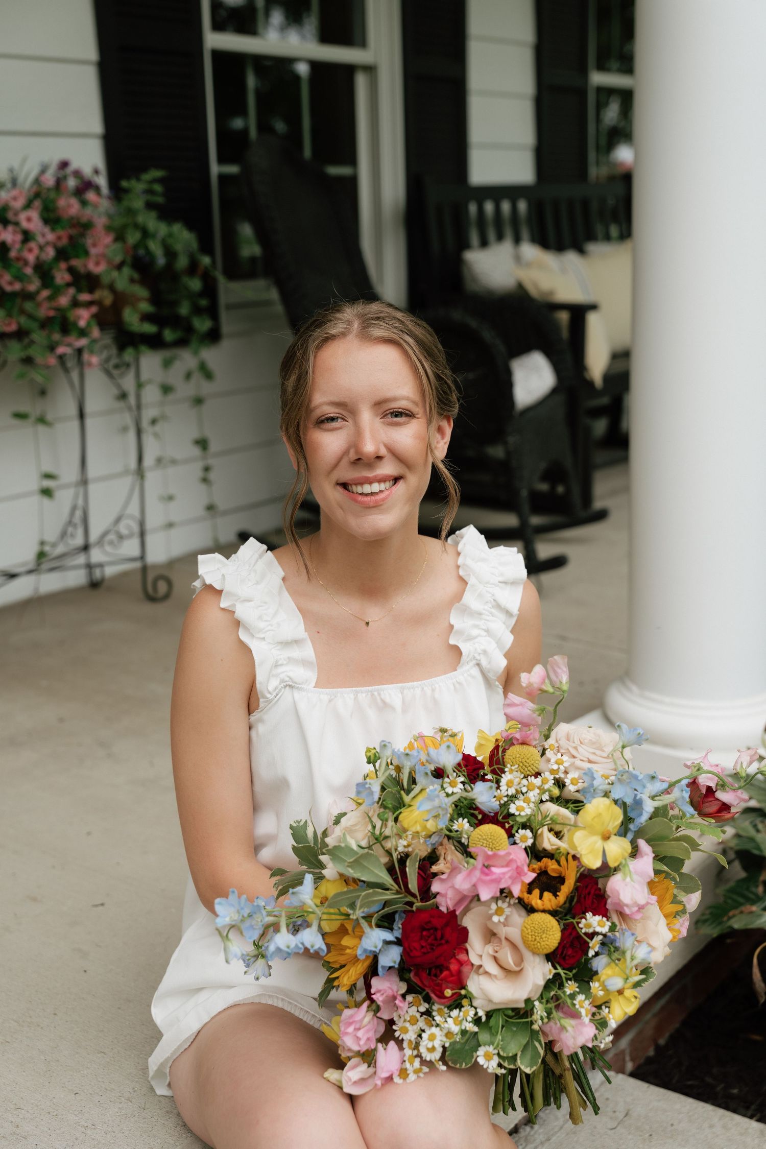 Smiling woman in white dress holding colorful wildflower bouquet on a porch.