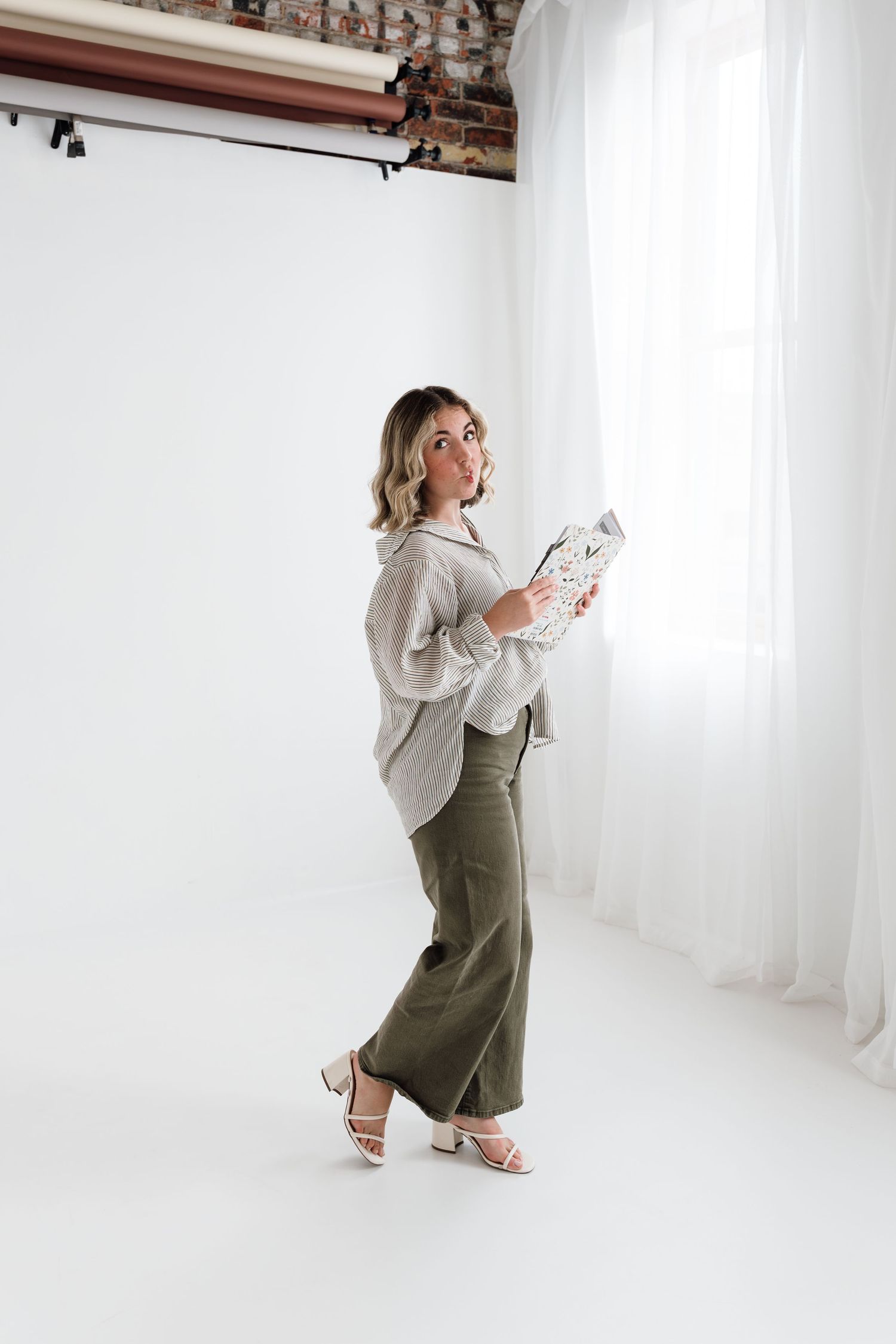 Woman in gray shirt and olive wide-leg pants holds magazine in bright white studio setting with sheer curtains.