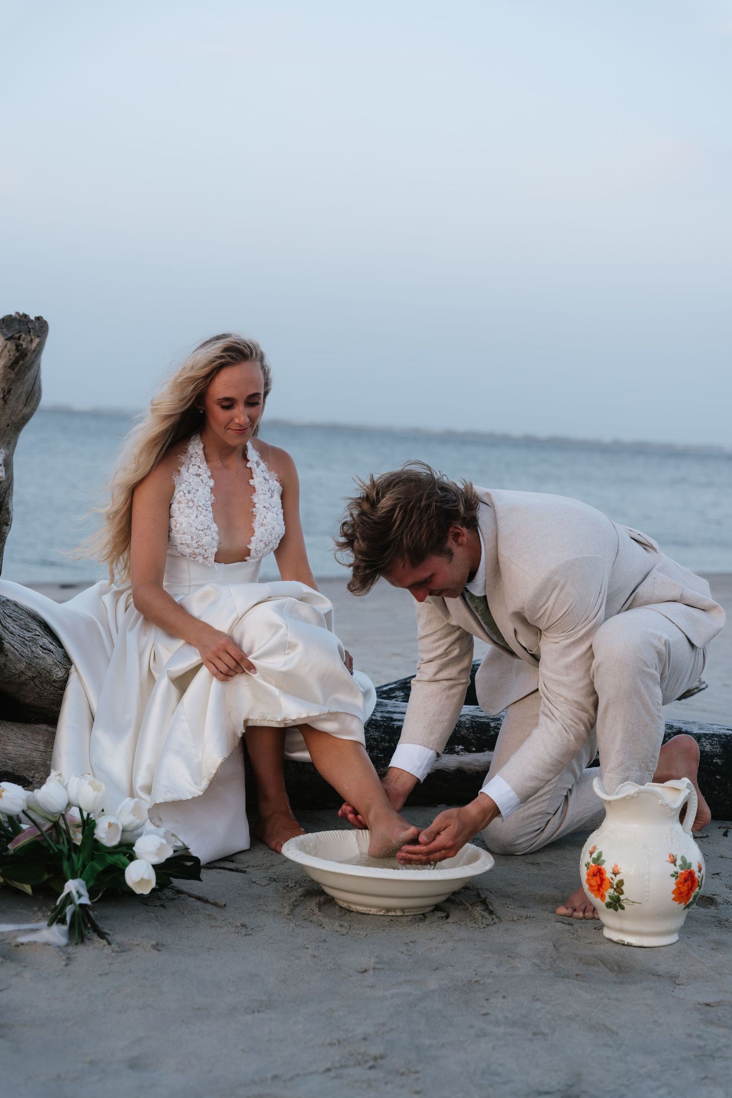 Groom washing bride's feet in a bowl on a sandy beach during a romantic outdoor wedding ceremony at dusk.