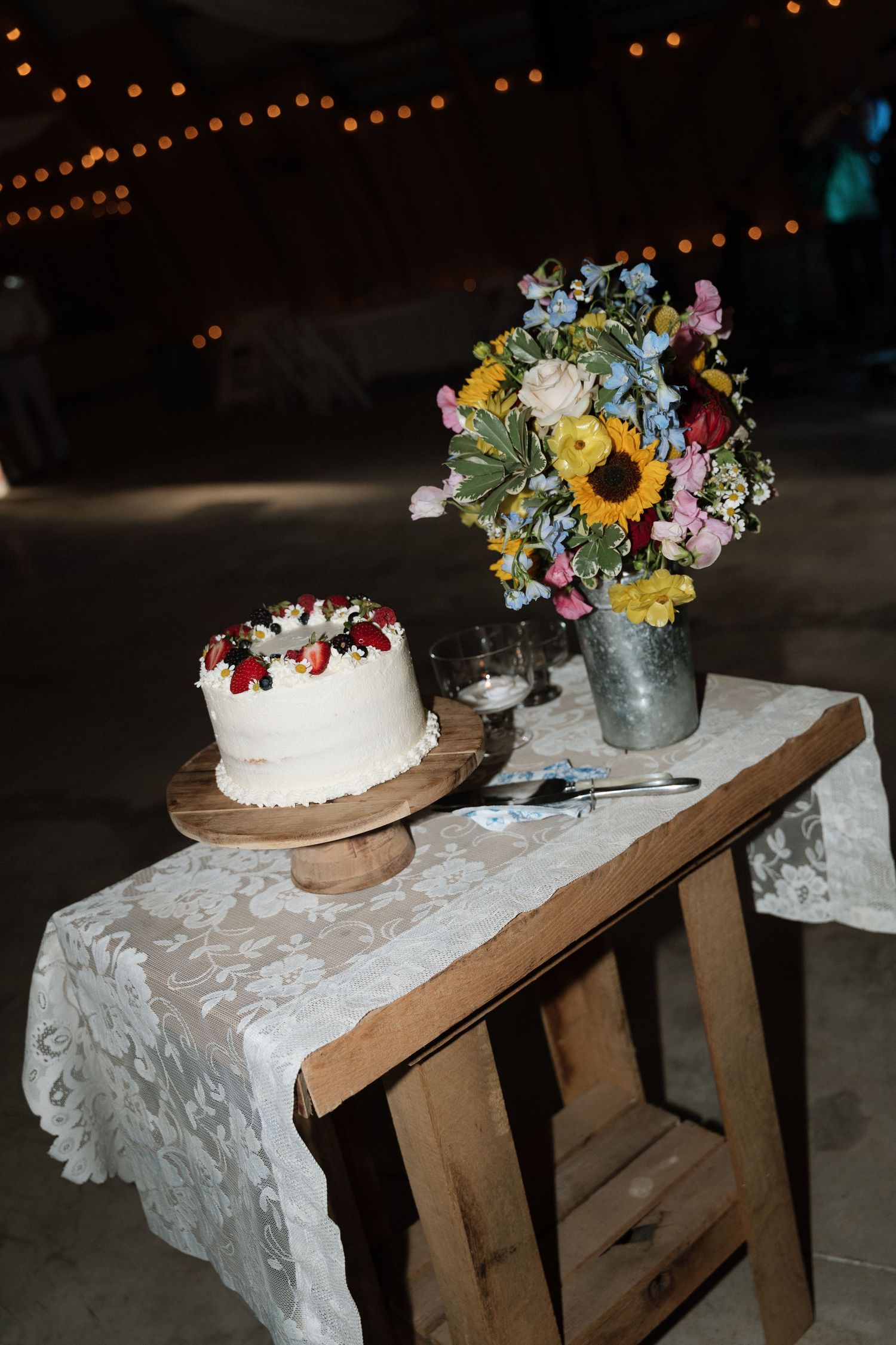 White wedding cake with berry topping on wooden stand beside wildflower bouquet with sunflowers on lace-covered rustic table.