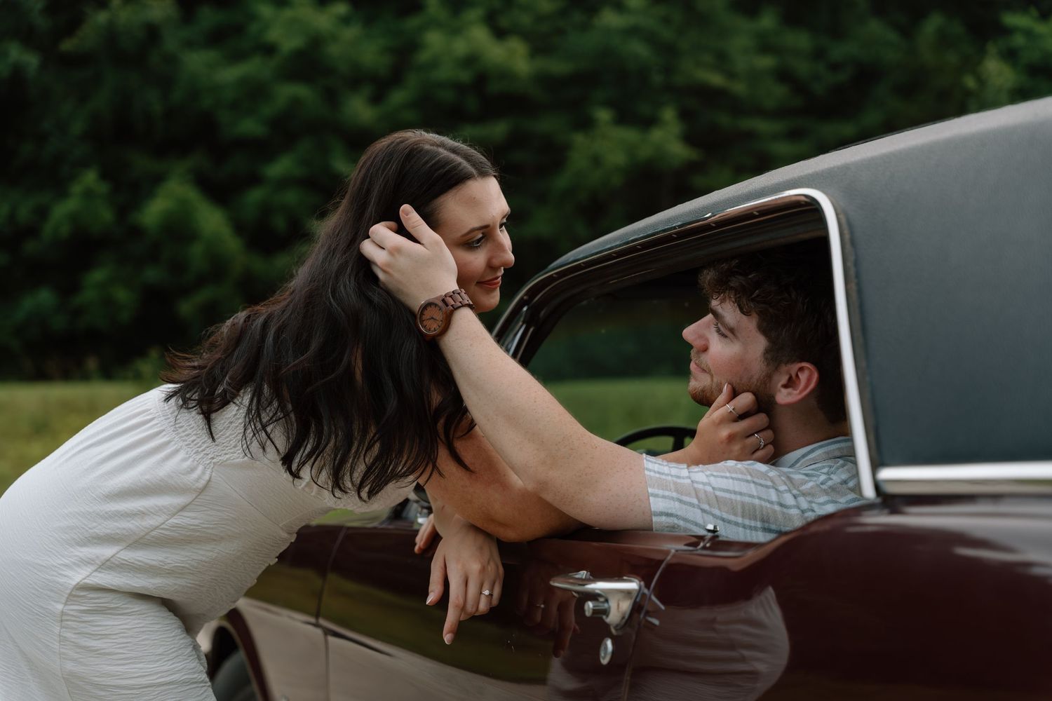 A woman leans into a vintage car window, tenderly holding a man's face in a romantic outdoor setting.