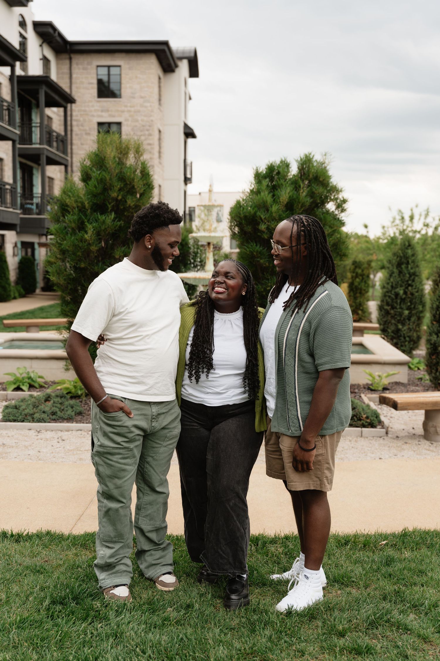Three friends smiling and laughing together outdoors near a fountain and green trees beside a modern building.
