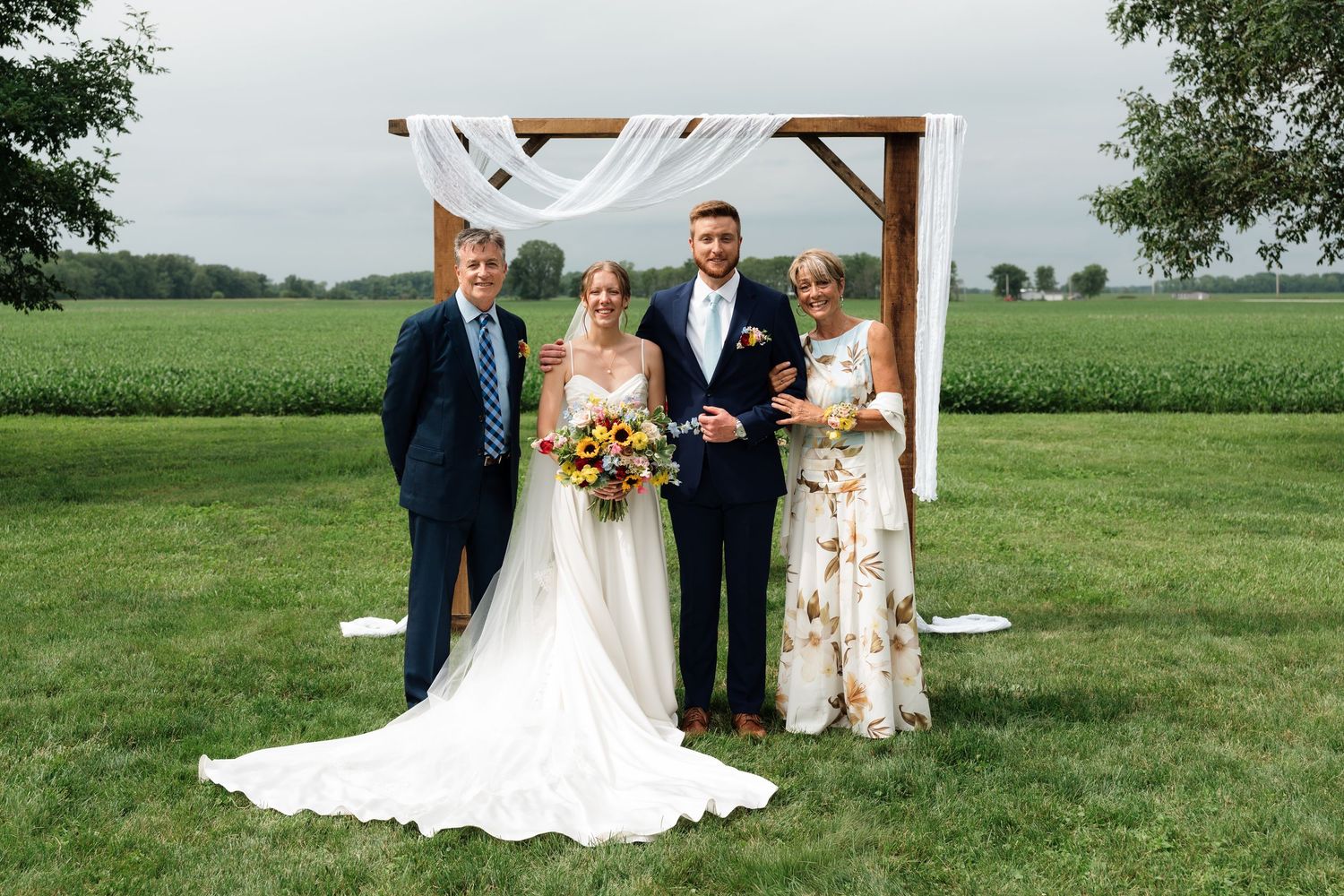 Wedding party poses under rustic wooden arch draped with white fabric in a lush green farm field setting.